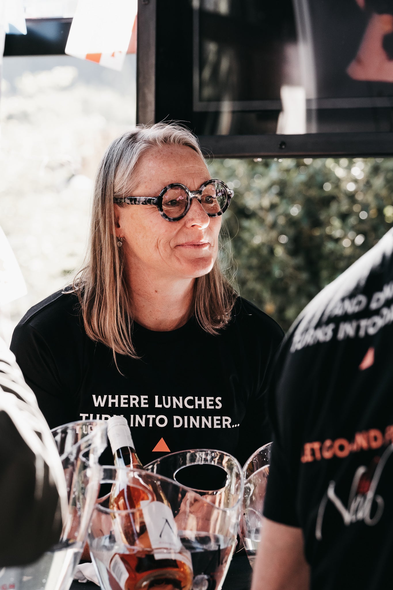 A person with long hair and glasses, wearing the mariscovip LET GO AND FEEL THE NED T-SHIRT, stands near a table laden with buckets of beverages. They appear to be at an outdoor event just before lunch, surrounded by sunlit greenery in the background.