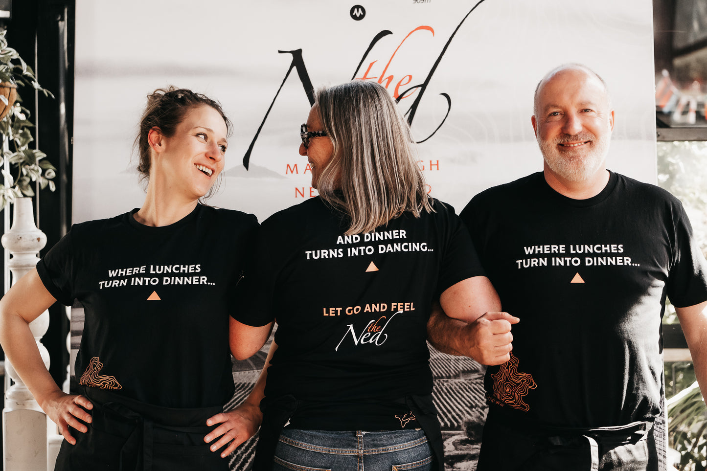 Three people smiling and standing with their backs to the camera, wearing LET GO AND FEEL THE NED T-SHIRTS by mariscovip, which feature text about dinners and dancing. A poster in the background adds context to the indoor setting.