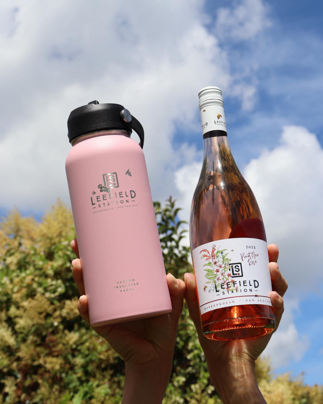 Two hands hold the LEEFIELD STATION DRINK BOTTLE - BLUSH, crafted from BPA-free and reusable stainless steel, next to a bottle of Marisco Family Vineyards' Leefield Station rosé wine, set against a backdrop of blue sky and lush green foliage.