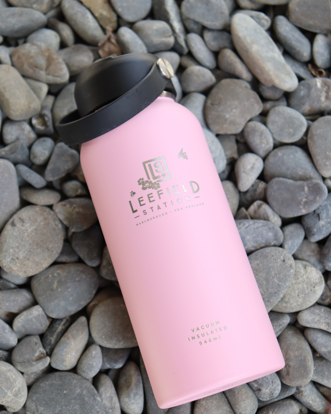 A blush-colored, reusable stainless steel vacuum insulated bottle with a black cap is resting on a bed of gray pebbles. This BPA-free bottle, named LEEFIELD STATION DRINK BOTTLE - BLUSH by Marisco Family Vineyards, features the text Leefield Station and Marlborough, New Zealand, along with 946ml.