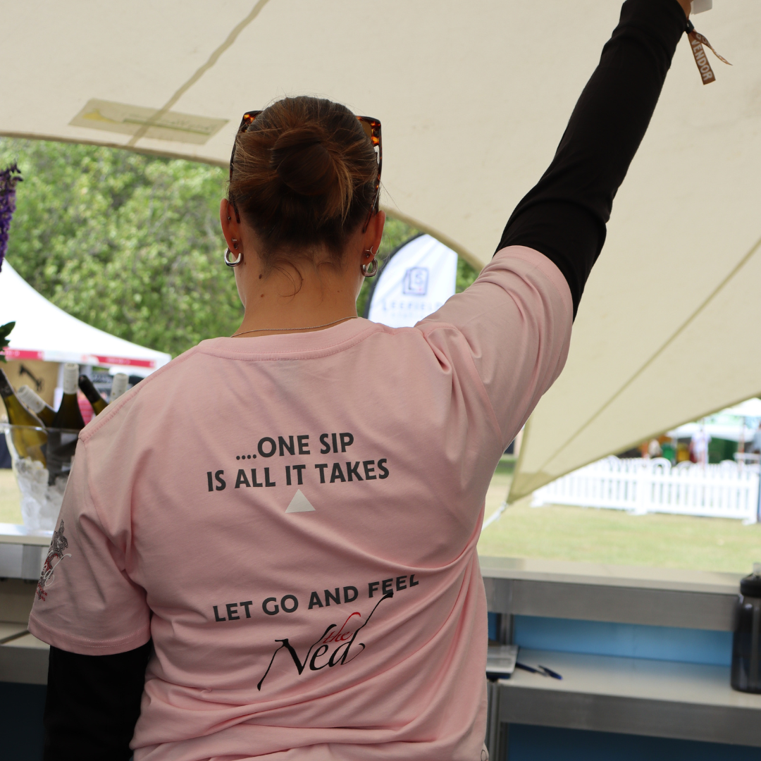 A person with their hair tied back and wearing sunglasses stands with an arm raised, creating a memorable moment. They're wearing a pink "CREATE THE MOMENT T-SHIRT" from Marisco Family Vineyards against the backdrop of an outdoor event.