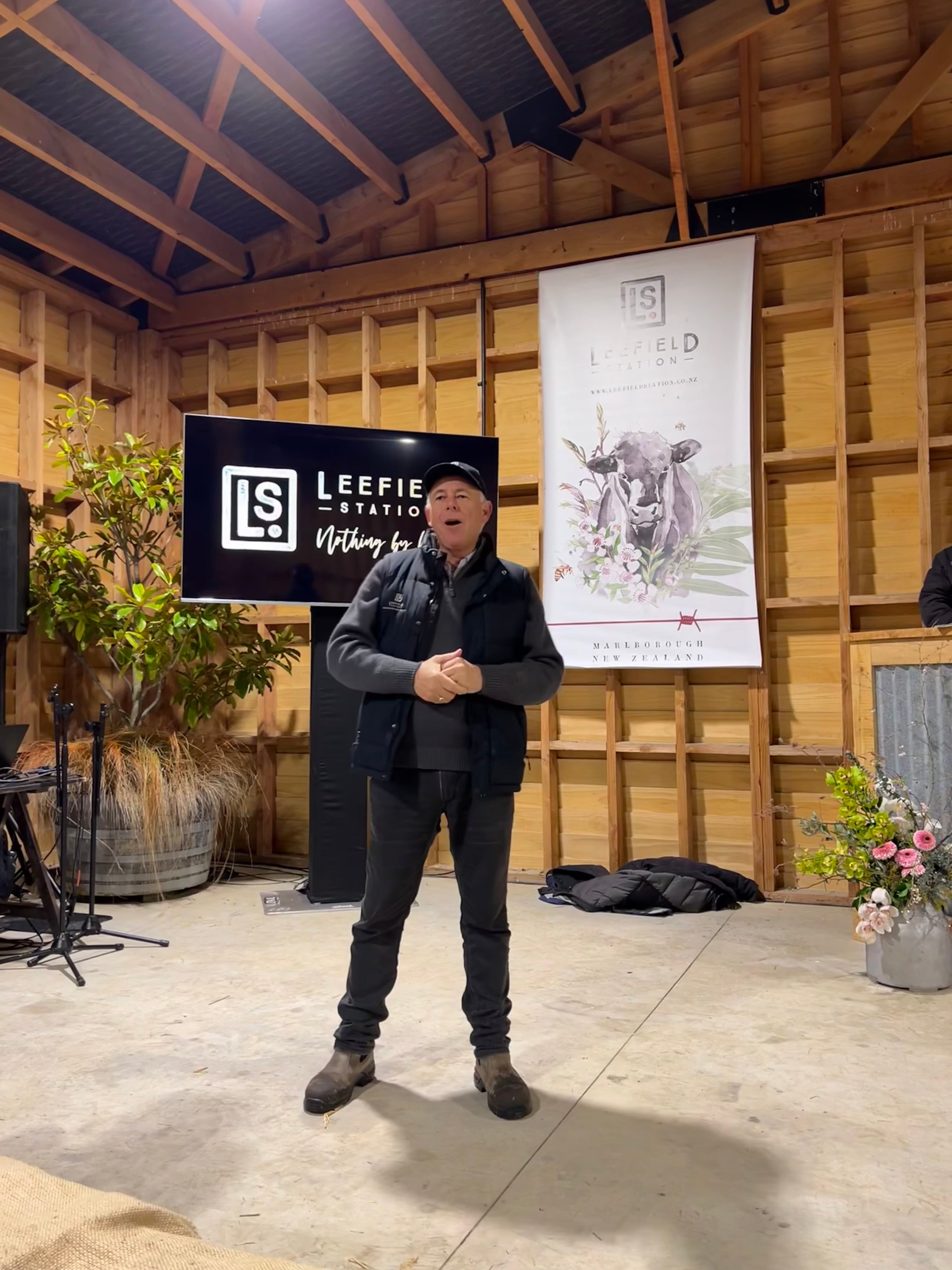 A person stands on a stage in a rustic wooden venue with a microphone. Behind them is a Leefield Station banner and a large display screen showing the same logo. A small decorative arrangement and plant are on the stage.