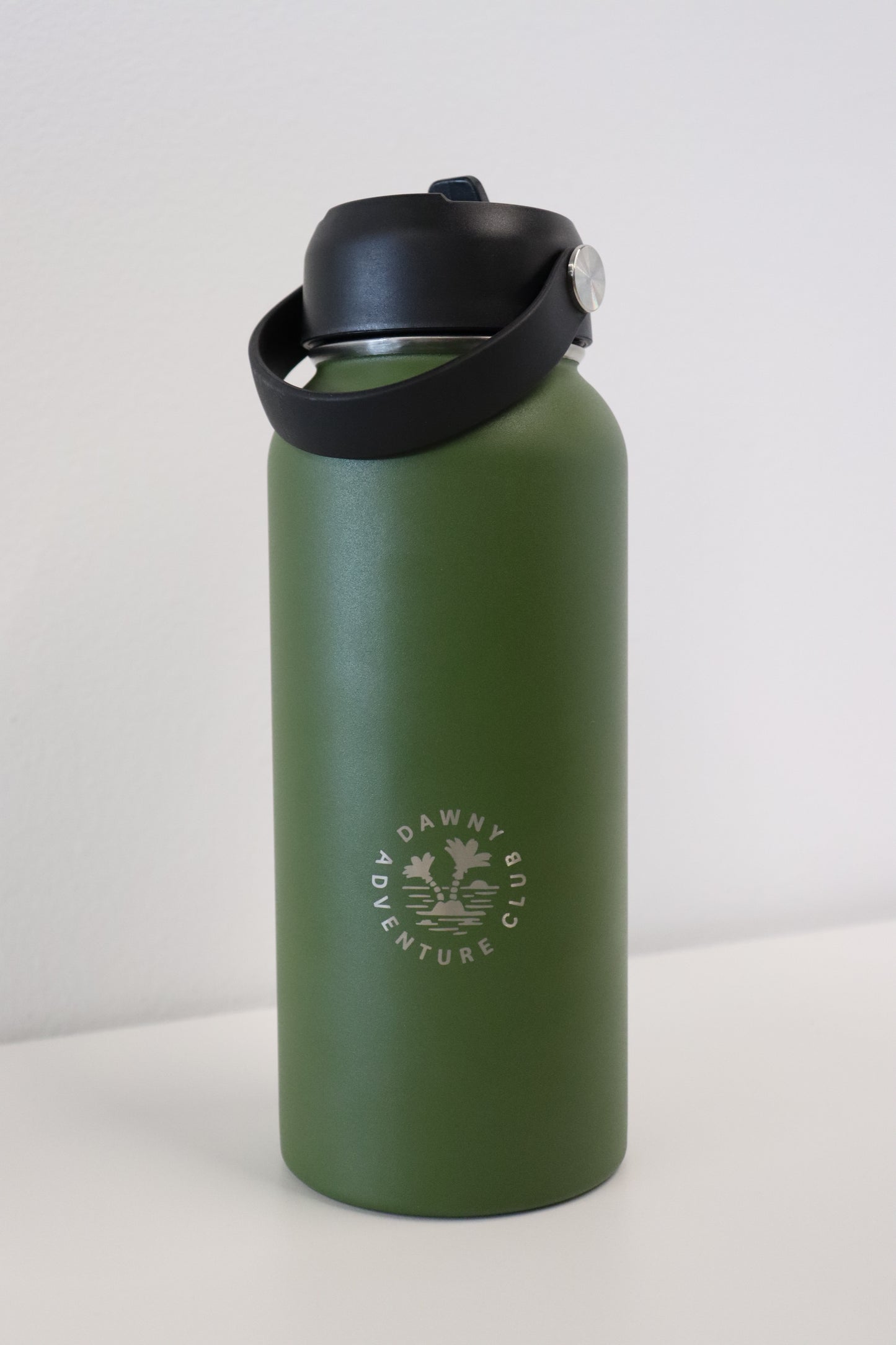 A green LEEFIELD STATION DRINK BOTTLE by Marisco Family Vineyards, featuring a black lid and handle. It is constructed from BPA-free stainless steel and includes dual-walled insulation. The logo, "Dawny Adventure Club," is displayed in white text. The bottle rests on a white surface against a light wall background.