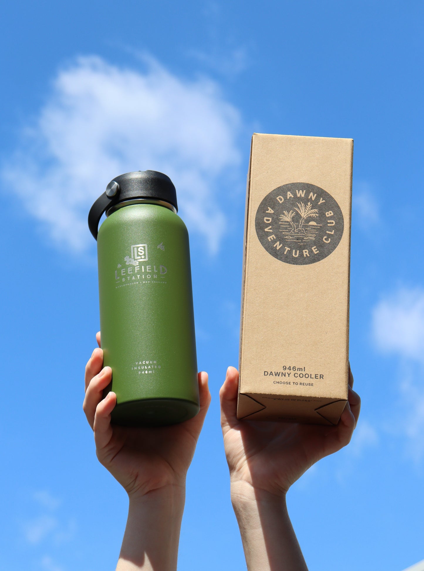 Two hands raise a green insulated water bottle, the LEEFIELD STATION DRINK BOTTLE by Marisco Family Vineyards, made from BPA-free, reusable stainless steel, alongside a brown cardboard box against a bright blue sky. The bottle features dual-walled insulation and a logo, while the box proudly reads Dawny Adventure Club.