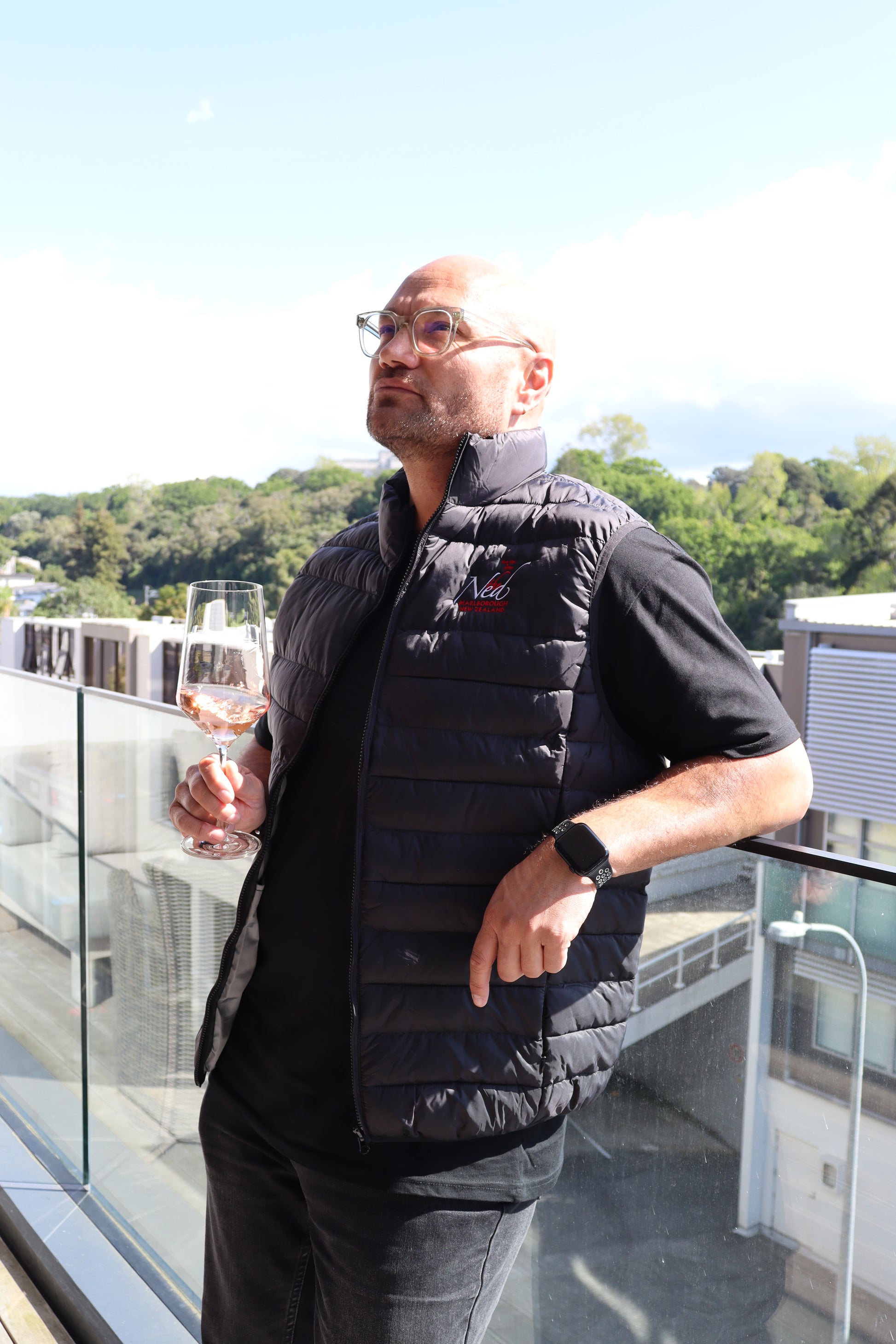 A man in glasses, a black shirt, and THE NED PUFFER VEST enjoys a glass of wine from Marisco Family Vineyards while standing on a balcony. He looks relaxed as he gazes into the distance, with trees and a clear sky in the background.