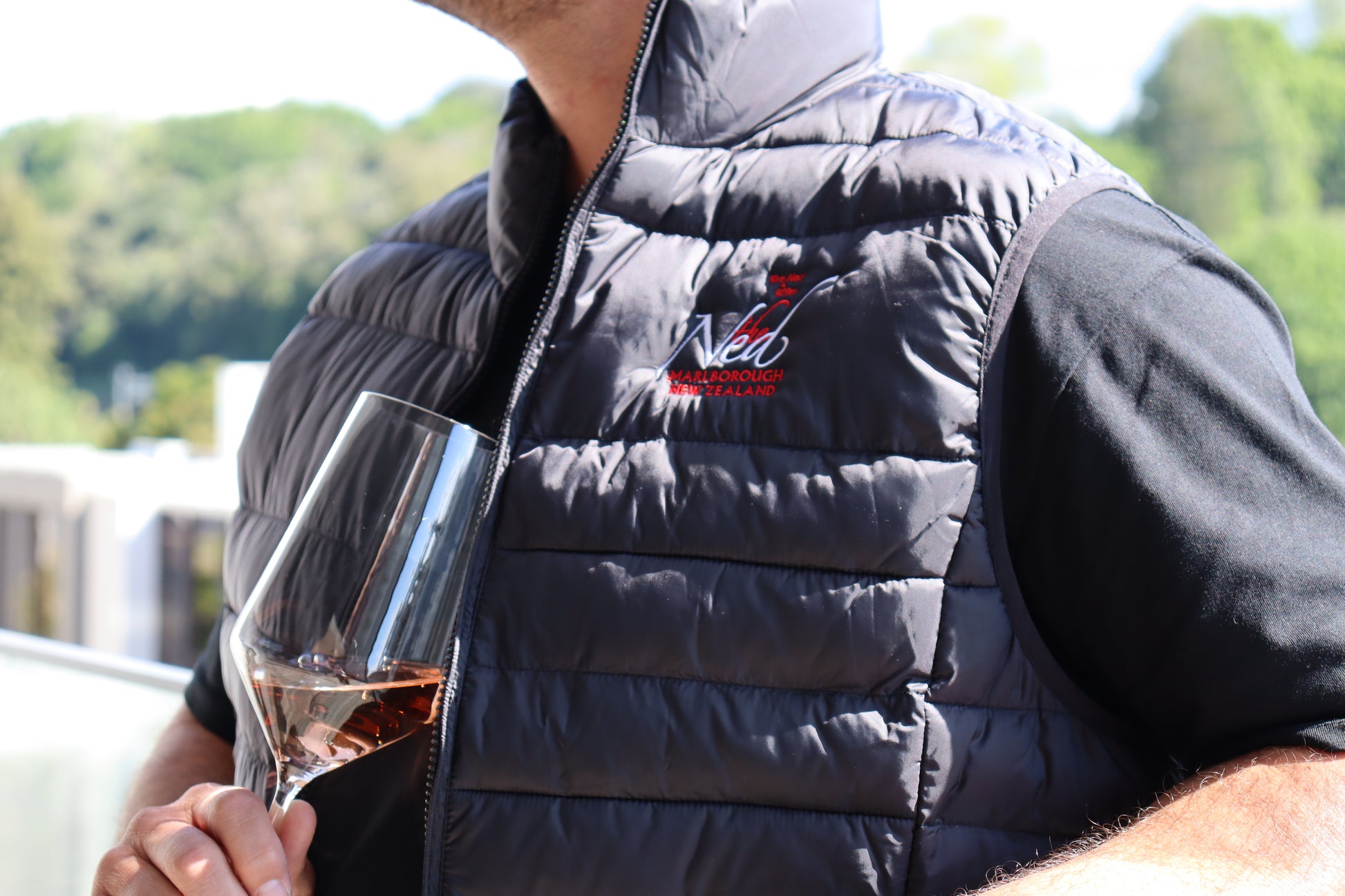 A person wearing THE NED PUFFER VEST with red embroidery holds a glass of Marisco Family Vineyards rosé wine. They are standing outdoors, and blurred greenery is visible in the background.