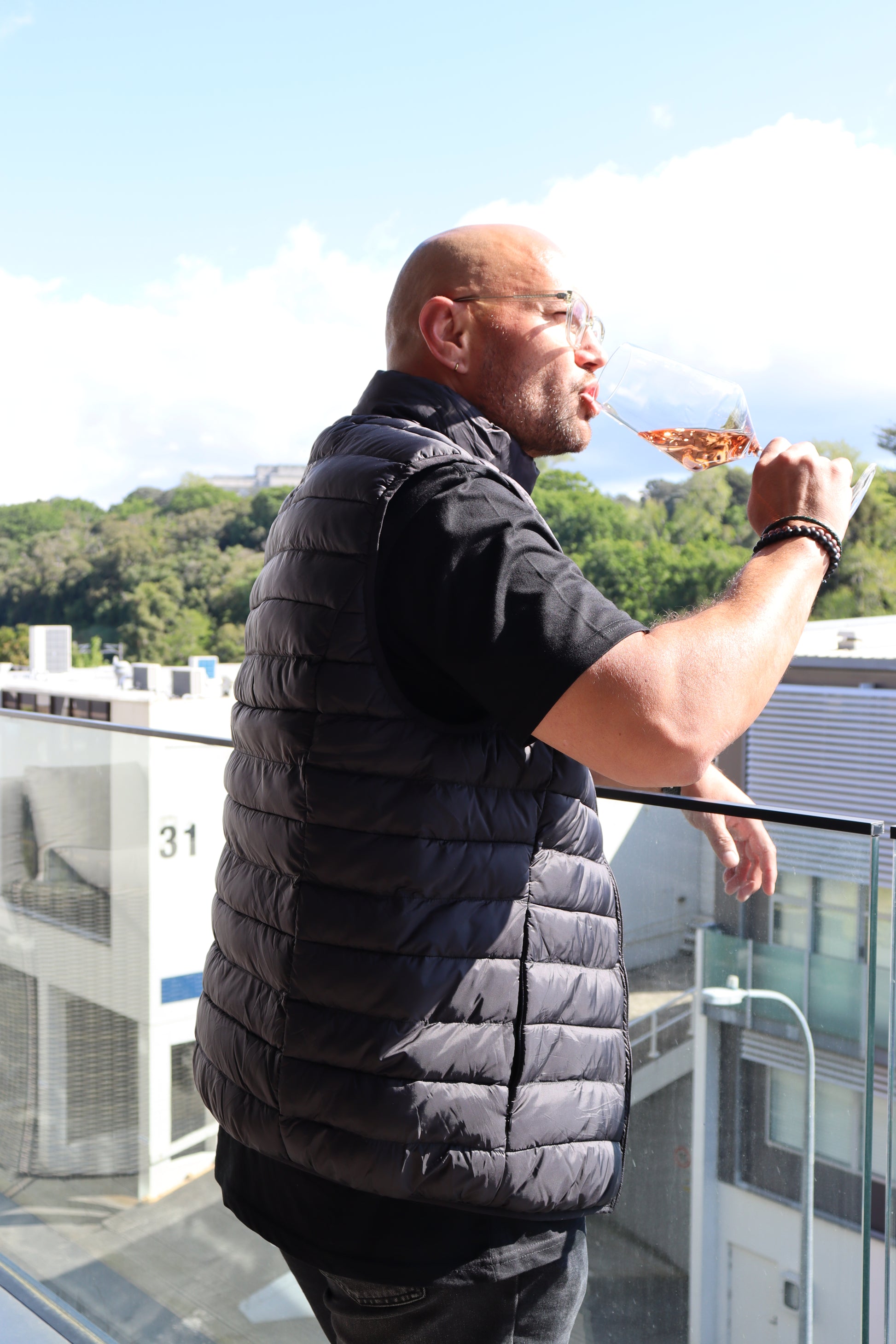 A man wearing THE NED PUFFER VEST enjoys a drink from a large glass while standing on a balcony overlooking lush green trees and a building with a blue and white facade, all illuminated by bright sunlight.