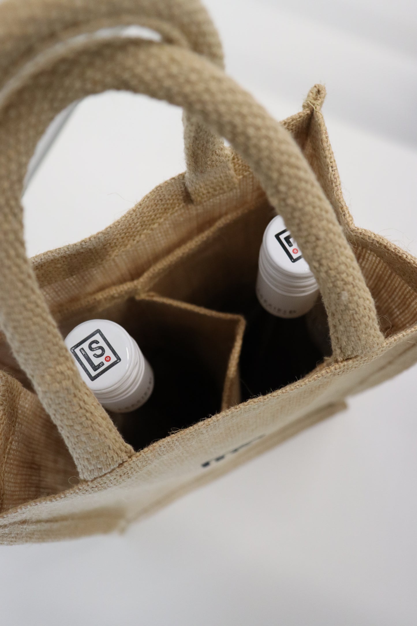 A LEEFIELD STATION TWO BOTTLE JUTE BAG by Marisco Family Vineyards, featuring a woven design, holds two bottles of wine with white caps. The bag's compartments and intricate material texture stand out against a neutral background when viewed from above.