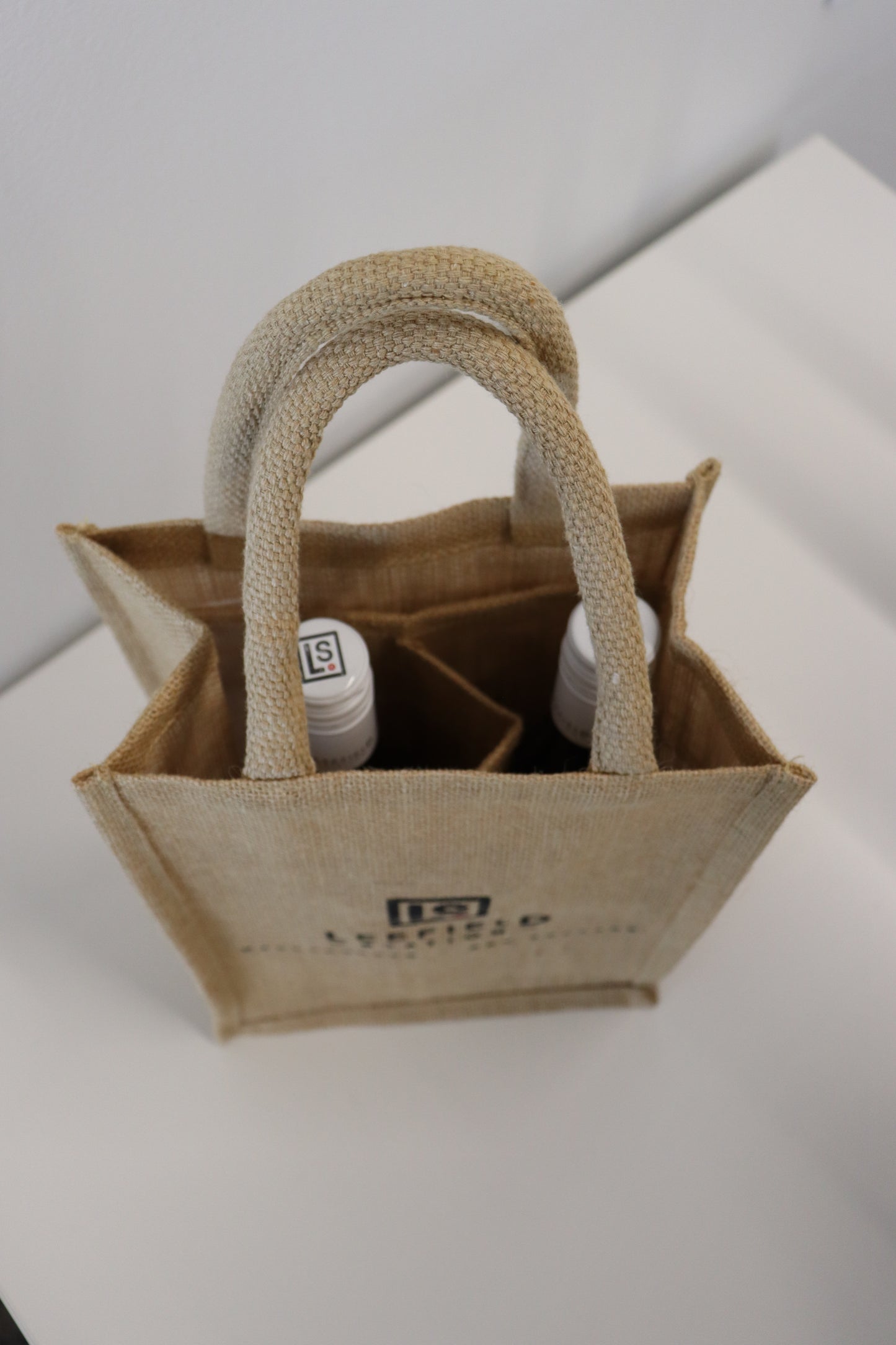 A top view of the LEEFIELD STATION TWO BOTTLE JUTE BAG from Marisco Family Vineyards, featuring two bottles nestled inside, rests on a white surface. The small beige jute gift bag showcases two handles and a stitched design, with part of a black logo visible on the front.