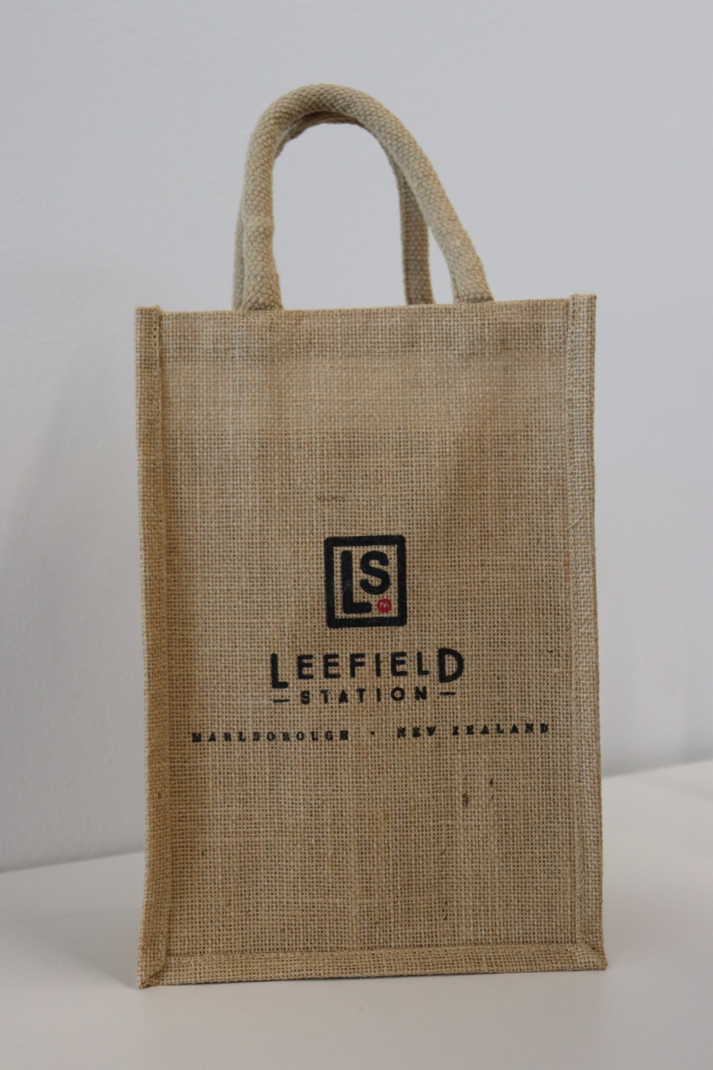 A chic LEEFIELD STATION TWO BOTTLE JUTE BAG with two handles rests against a white backdrop, evoking the style of a sophisticated gift bag. The beige jute bag features a logo showcasing the initials LS along with "LEEFIELD STATION" and "MARLBOROUGH, NEW ZEALAND" printed in black. This elegant accessory from Marisco Family Vineyards is ideal for carrying or gifting two bottles of Leefield Station Wines to someone special.