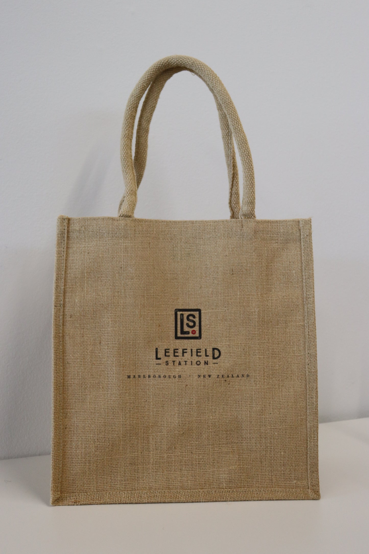 Introducing the LEEFIELD STATION LARGE JUTE BAG by Marisco Family Vineyards: This sturdy jute shopping bag comes in a natural color and features robust handles. The front is adorned with a logo, displaying 'Leefield Station' and 'Marlborough, New Zealand' in dark brown text. It’s an ideal choice for carrying groceries, photographed against a simple white background.