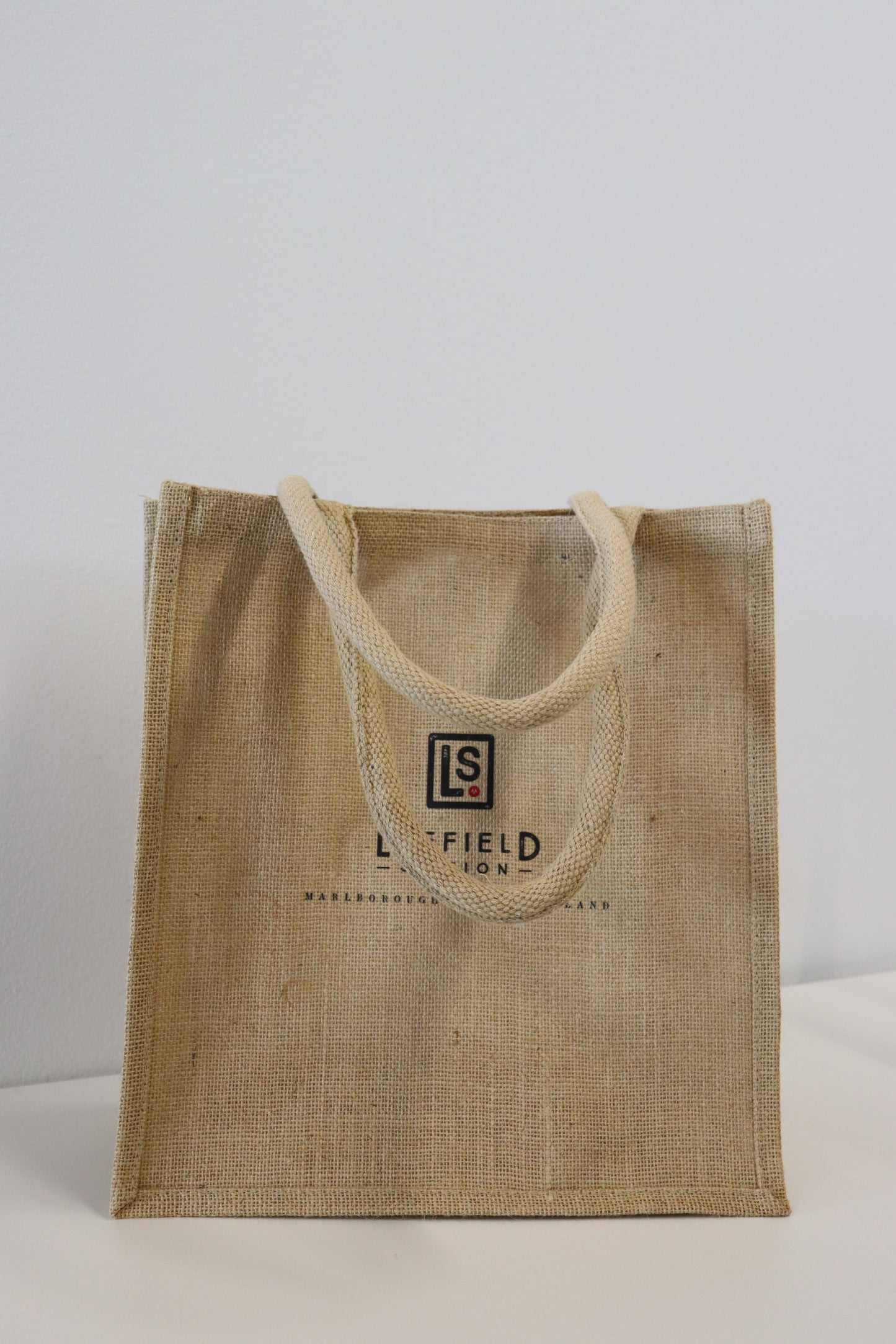 A tan jute tote bag from Marisco Family Vineyards, known as the LEEFIELD STATION LARGE JUTE BAG, rests on a white surface. Designed with two handles, it features "LS" and "L. Field" text on the front. The simple design stands out against a plain white wall backdrop, making it an ideal eco-friendly grocery companion.