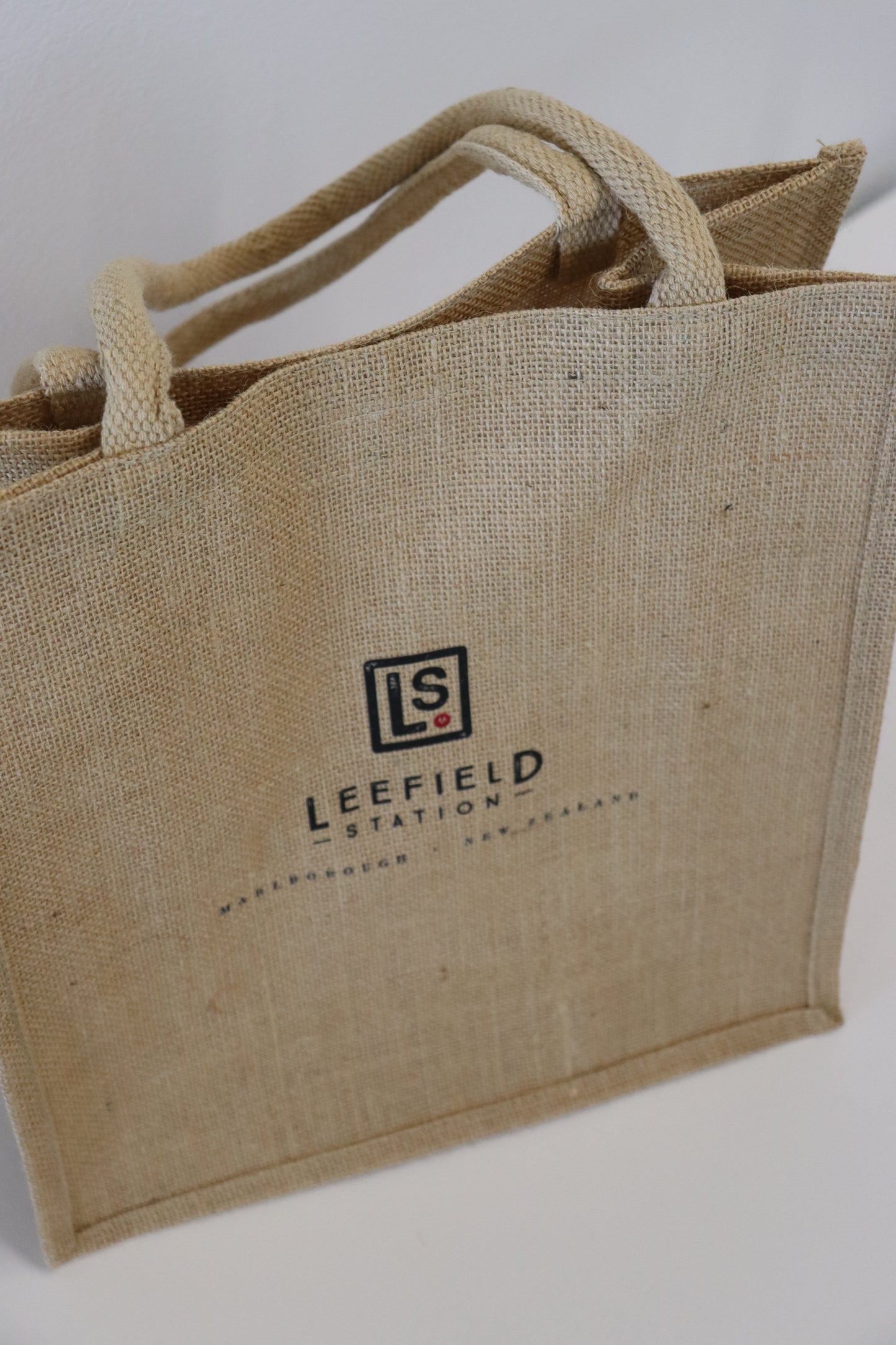 The LEEFIELD STATION LARGE JUTE BAG by Marisco Family Vineyards is a beige tote made of woven jute, featuring the LS logo and the words "Leefield Station Marlborough New Zealand." Ideal for shopping trips, it has strong, short handles and is displayed against a white background.