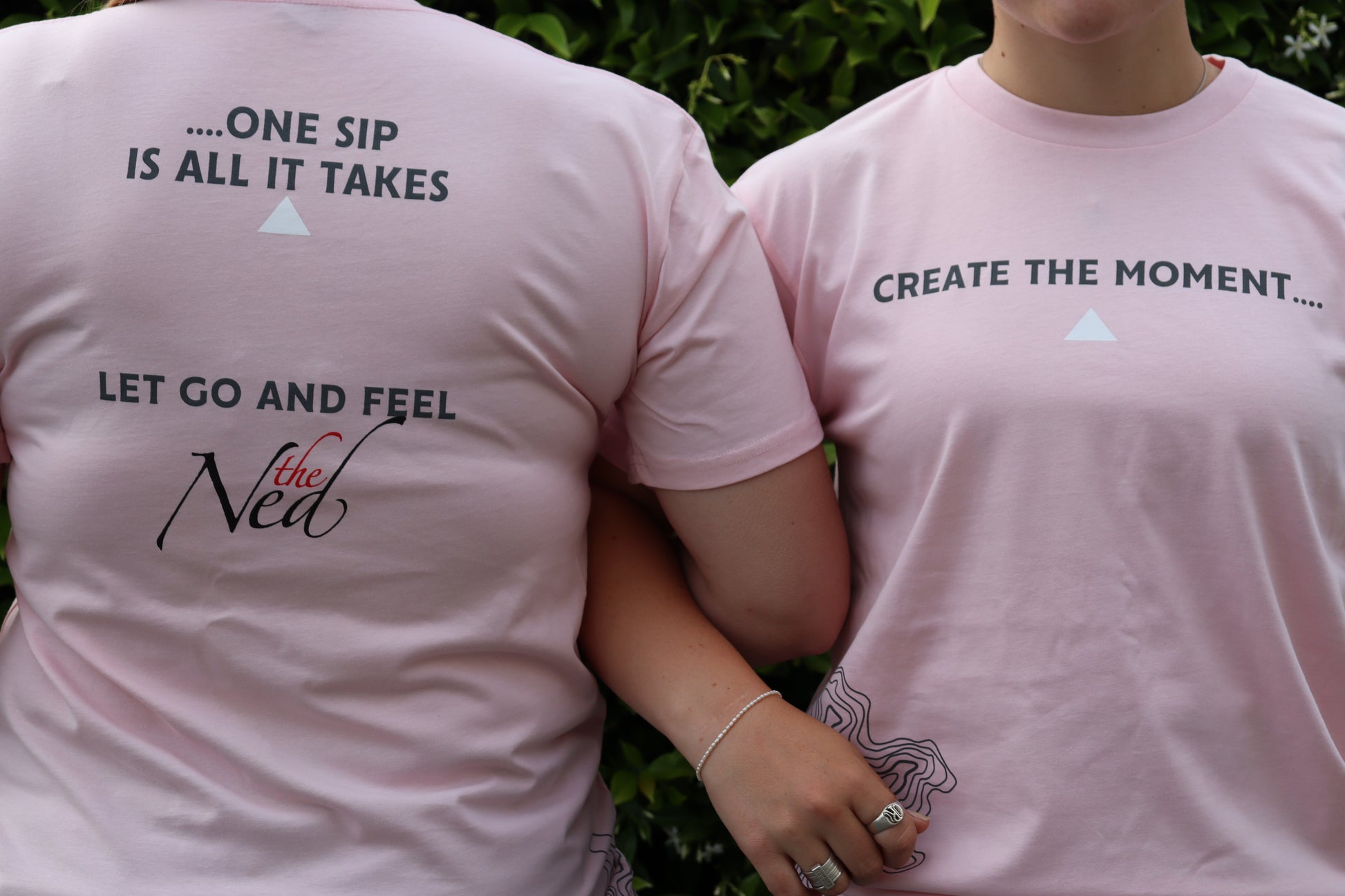 Two individuals are outdoors amidst lush greenery, wearing CREATE THE MOMENT T-SHIRTS in light pink by Marisco Family Vineyards. The back of the shirts features phrases like "...one sip is all it takes" and "let go and feel the Ned," while the front bears the slogan "create the moment.
