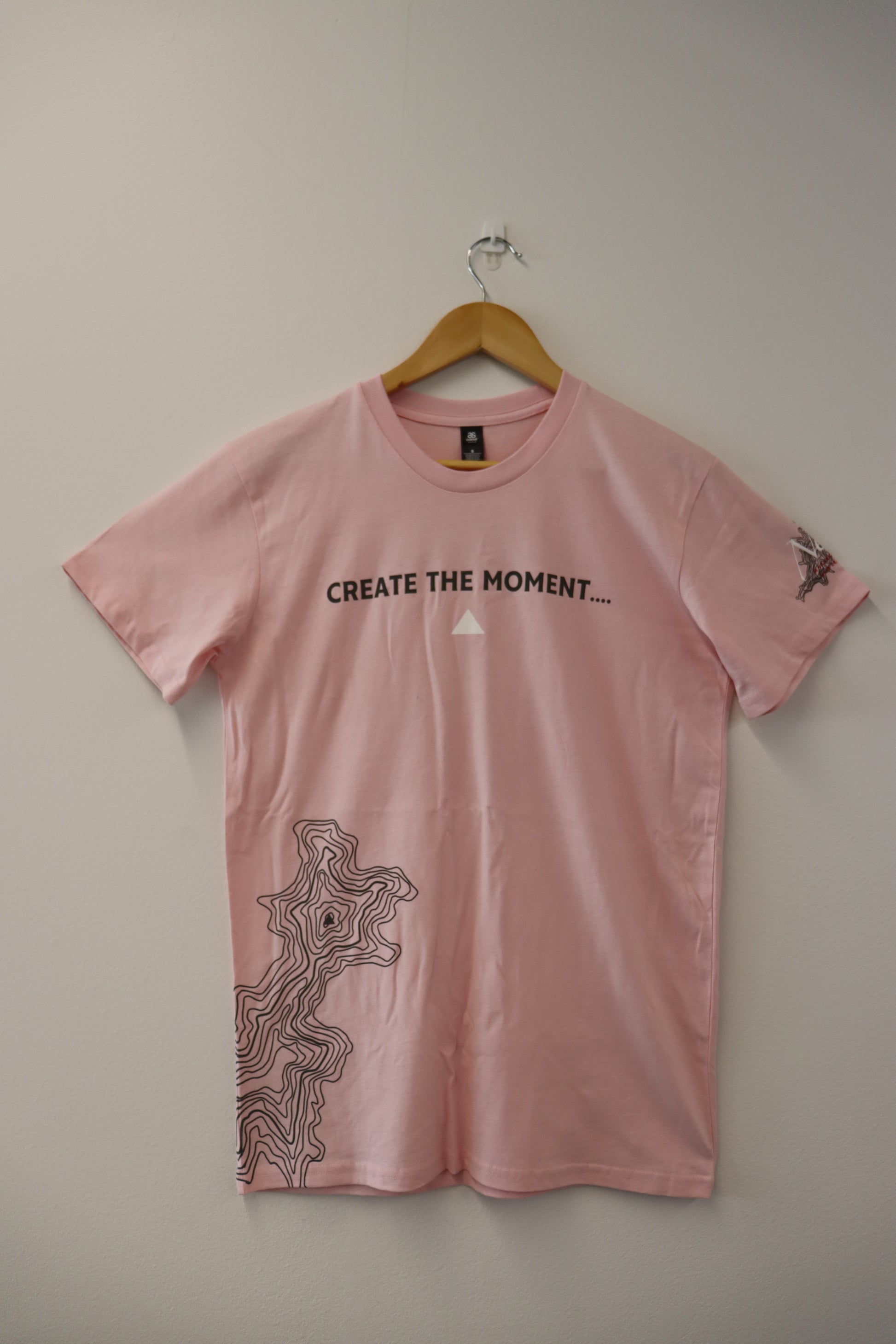 The CREATE THE MOMENT T-SHIRT by Marisco Family Vineyards is a pink tee boldly showcasing "CREATE THE MOMENT..." across the front. It features an abstract line pattern on the lower side, ideal for making a statement as you enjoy your favorite drink. Displayed on a wooden hanger against a plain wall, it embodies simplicity at its finest.