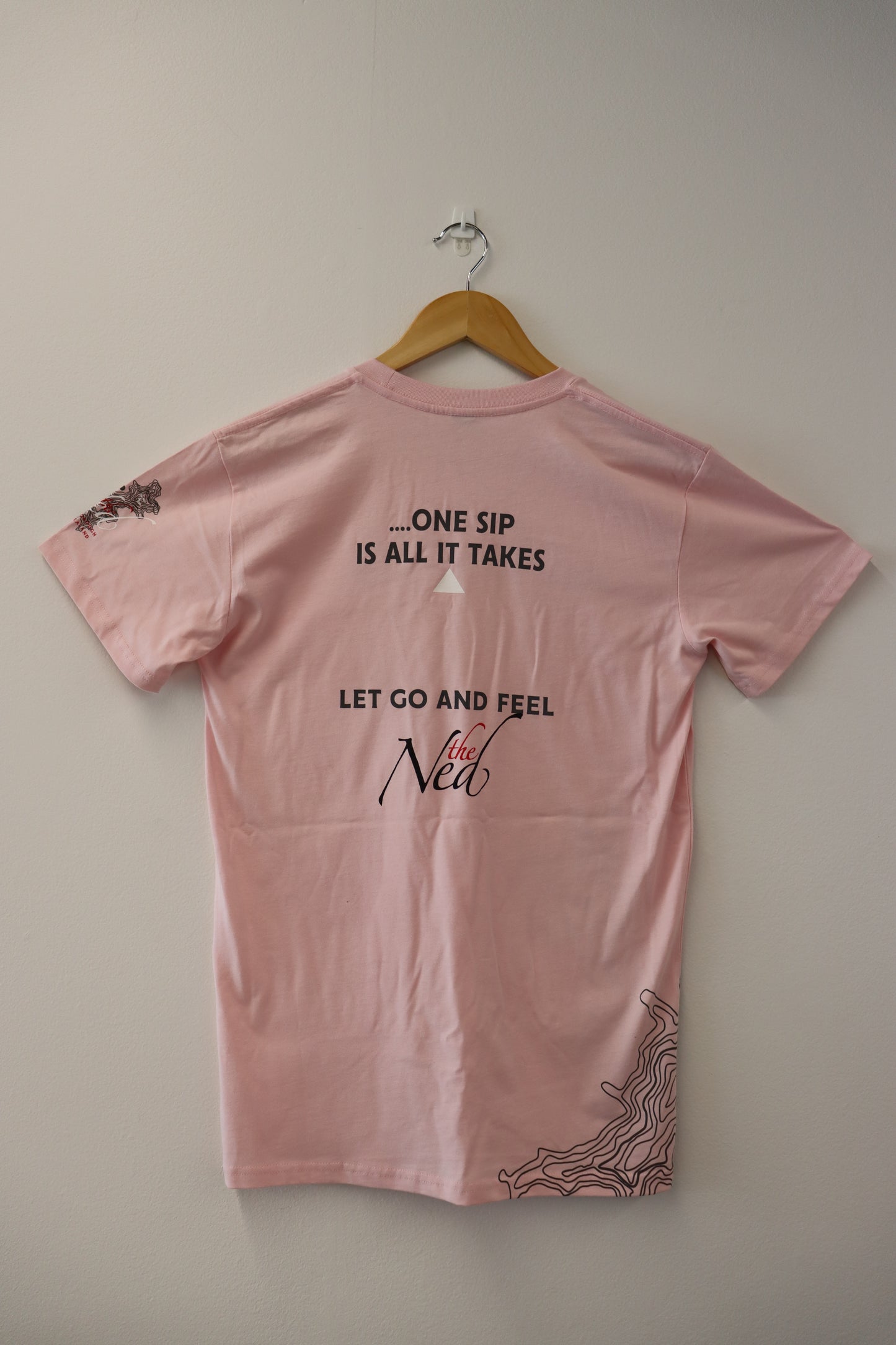 A pink CREATE THE MOMENT T-SHIRT by Marisco Family Vineyards, suspended on a wooden hanger against a white wall, beckons you with its messages: ONE SIP IS ALL IT TAKES and LET GO AND FEEL the NED. The sleeves and hem feature abstract line art, lending an artistic flair.