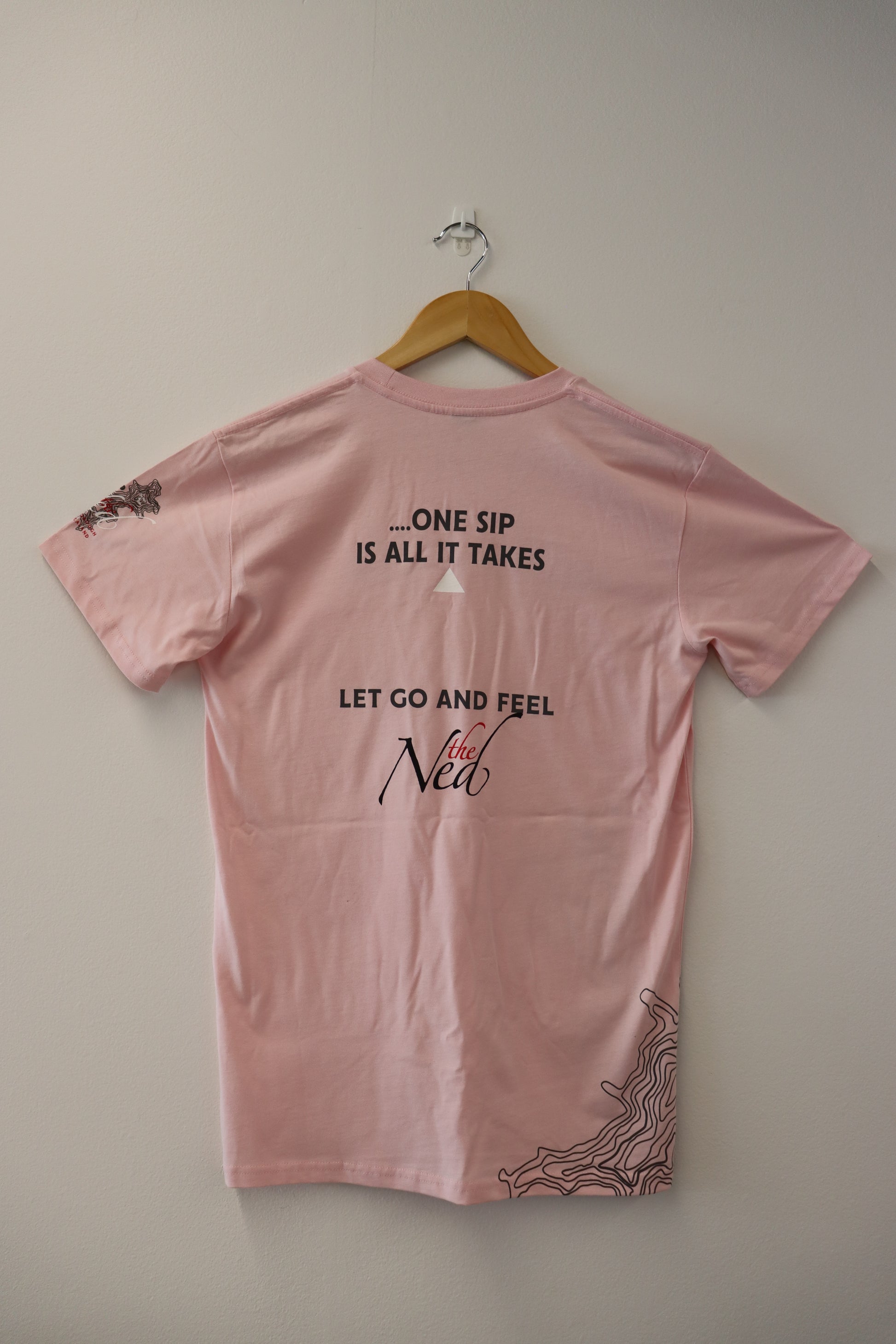 A pink CREATE THE MOMENT T-SHIRT by Marisco Family Vineyards, suspended on a wooden hanger against a white wall, beckons you with its messages: ONE SIP IS ALL IT TAKES and LET GO AND FEEL the NED. The sleeves and hem feature abstract line art, lending an artistic flair.