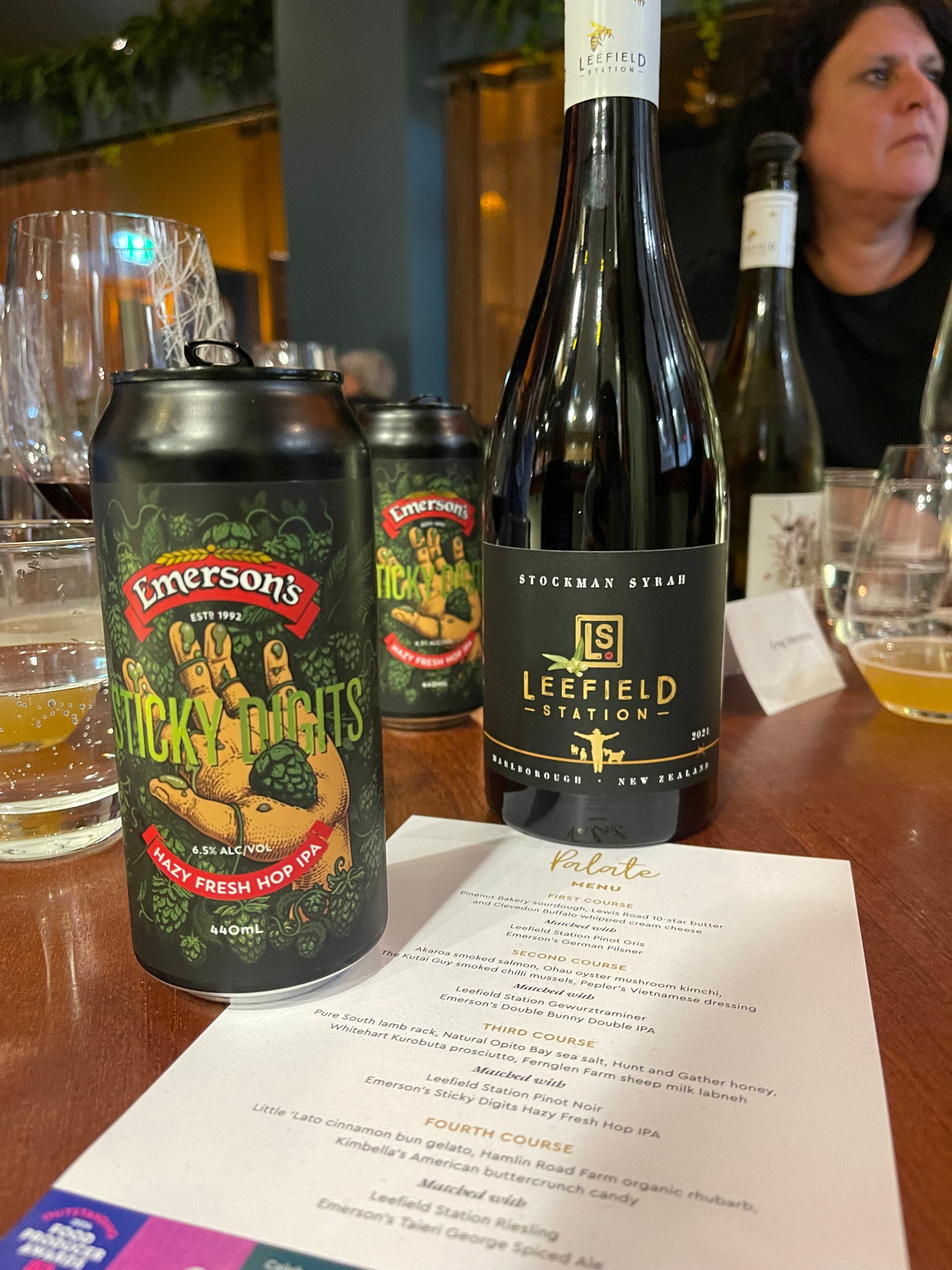 A table at a restaurant with a can of Emersons Sticky Digits hazy fresh hop IPA and a bottle of Leefield Station Stockmans Syrah. There are glasses and a menu listing different courses with beer pairings. People are seated around the table.