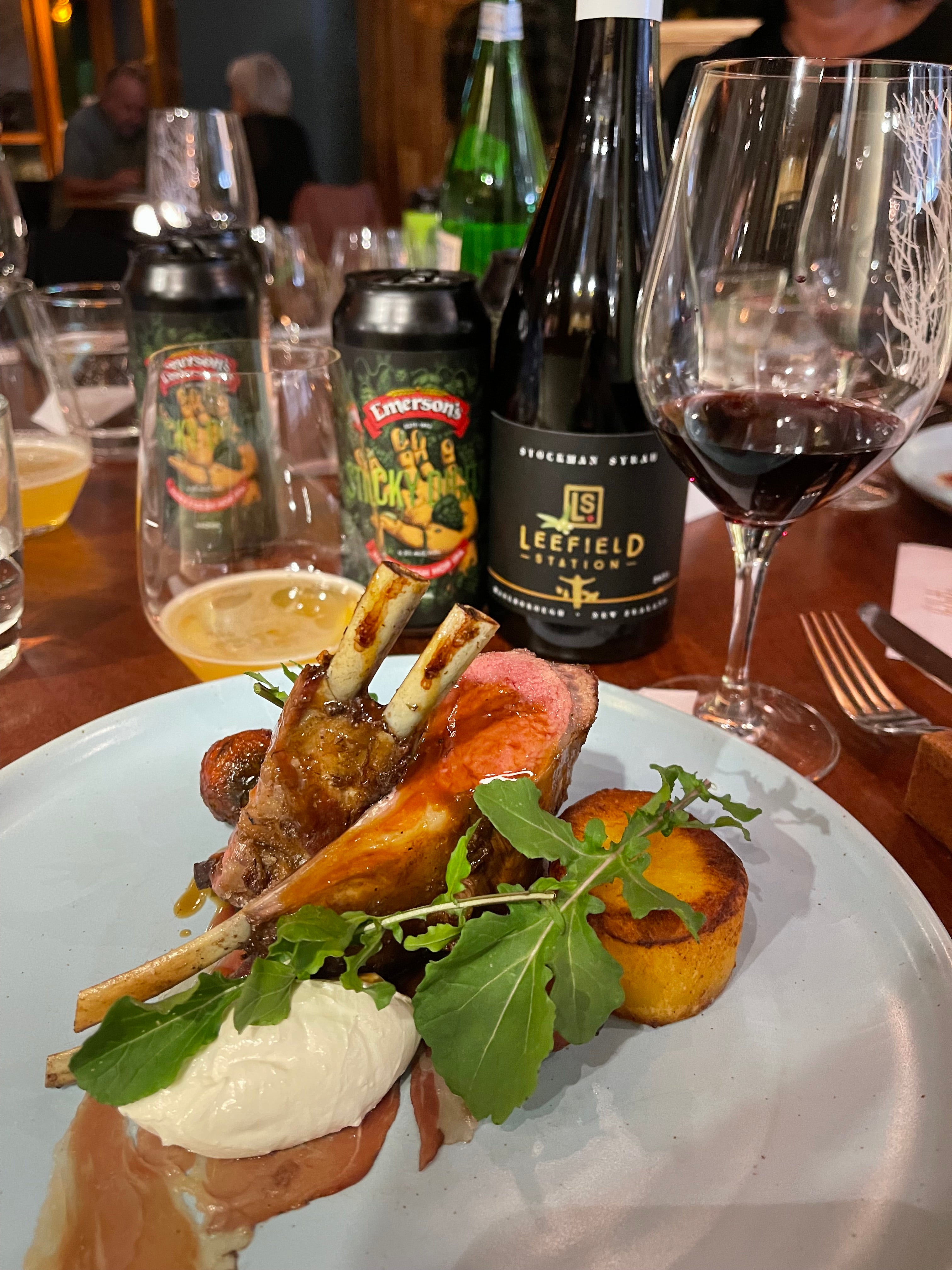 A plate of lamb chops garnished with arugula, served with a round potato cake and a dollop of cream. In the background, there are glasses of beer, a bottle of red wine, and wine glasses on a wooden table.
