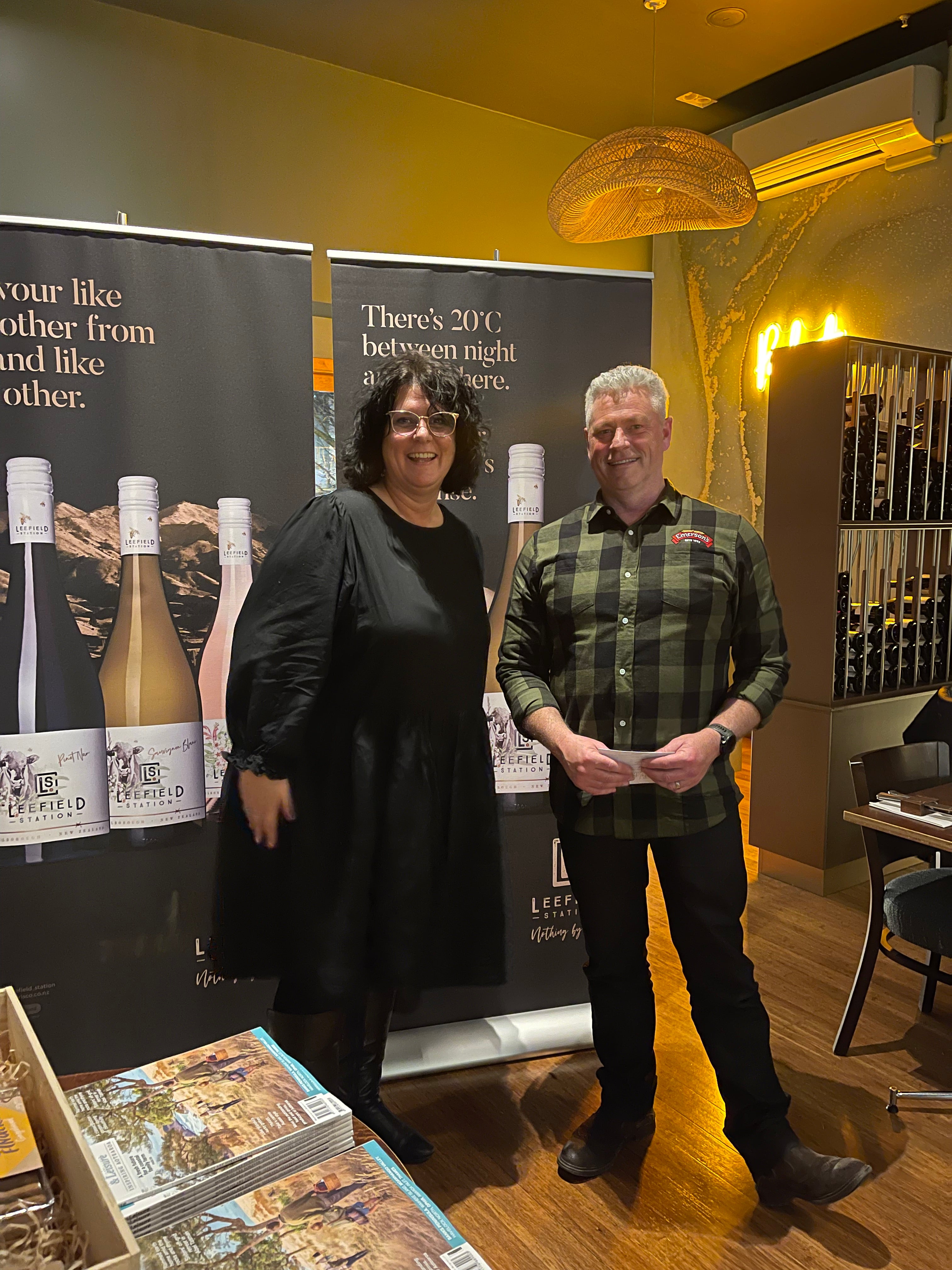 A woman and a man stand smiling in front of Leefield wine banners inside a warmly lit room. The setting appears to be a wine event or tasting, with wine bottles and promotional materials visible nearby.