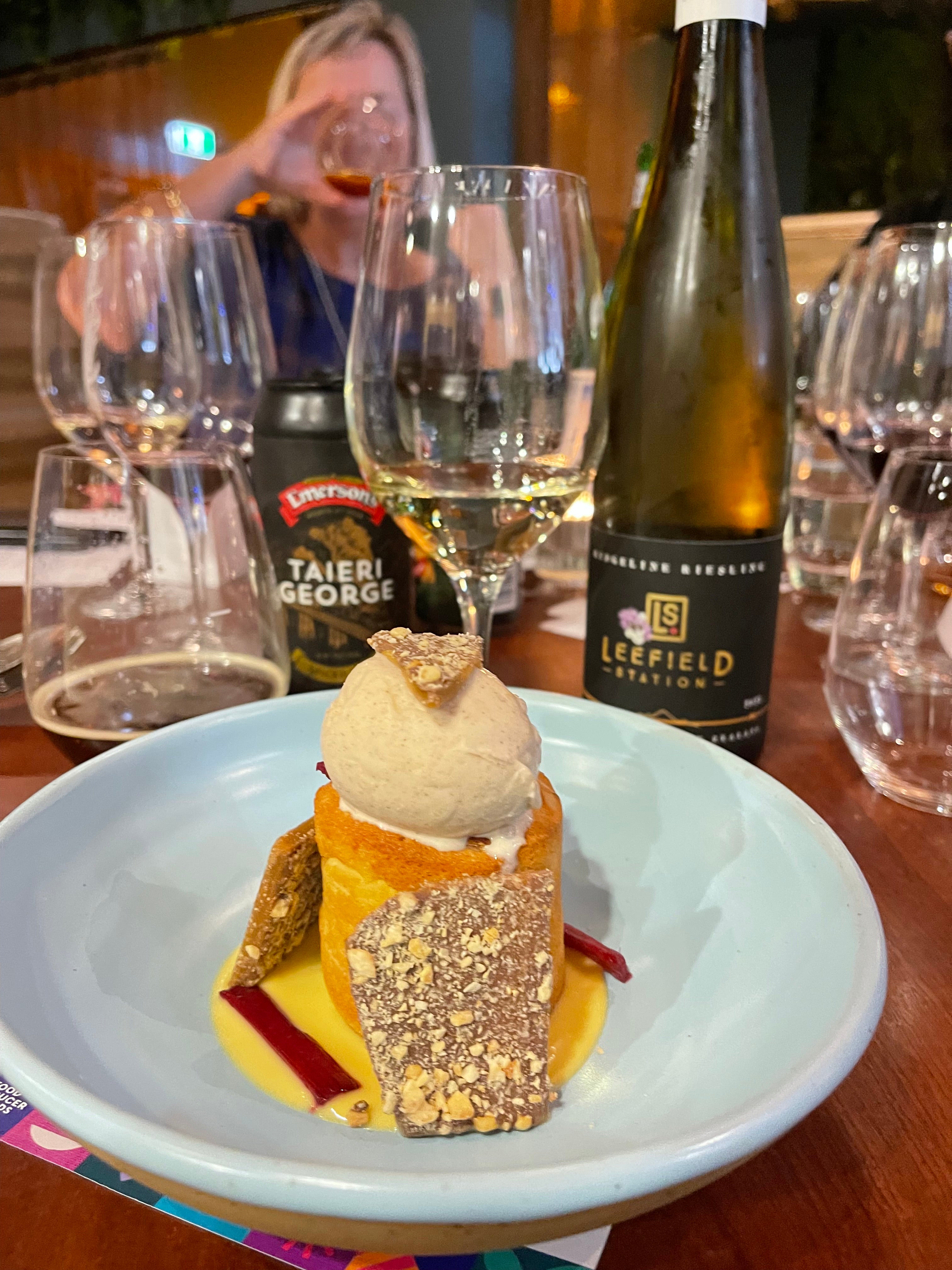 A dessert with ice cream topped on a pastry, garnished with sauce and brittle, sits on a plate. There are glasses and bottles of wine and beer on the table. A person is visible in the blurred background.