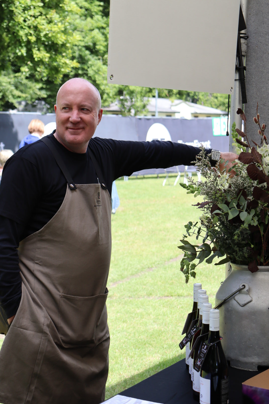 A man wearing the LEEFIELD STATION APRON from Marisco Family Vineyards stands at an outdoor booth with wine bottles arranged on a table. He smiles and gestures towards a plant arrangement in a vase, with a grassy area and people walking in the background.