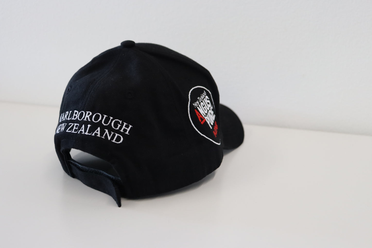 The LEEFIELD STATION CAP from Marisco Family Vineyards is placed on a white surface. The back of the black cap showcases "Marlborough New Zealand" in white stitching, while the side features a circular logo with red and white accents.