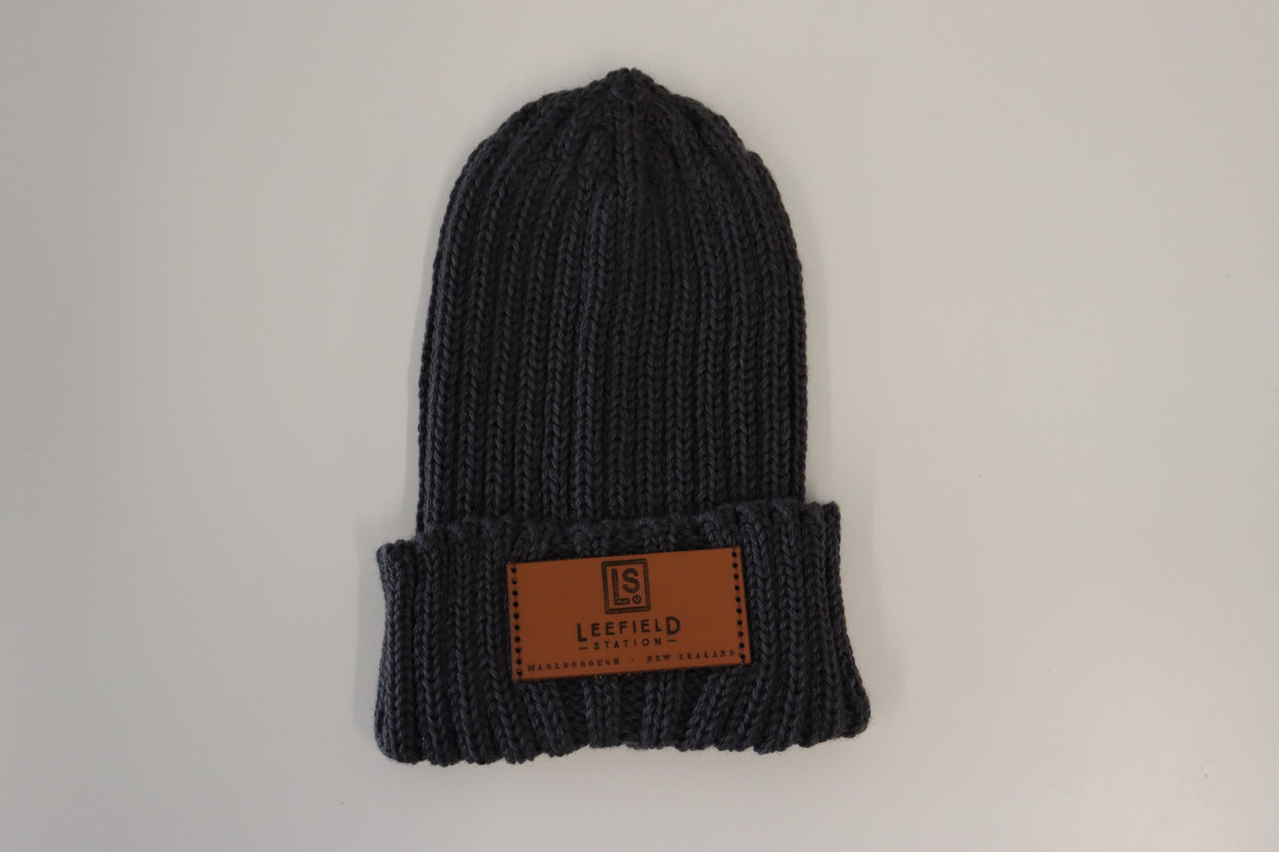 A flat-laid LEEFIELD STATION BEANIE from Marisco Family Vineyards, made of dark grey knit fabric with a folded cuff. It features a brown leather patch embossed with a logo and the text "LeeField - Established, Made in New Zealand," against a simple light background.