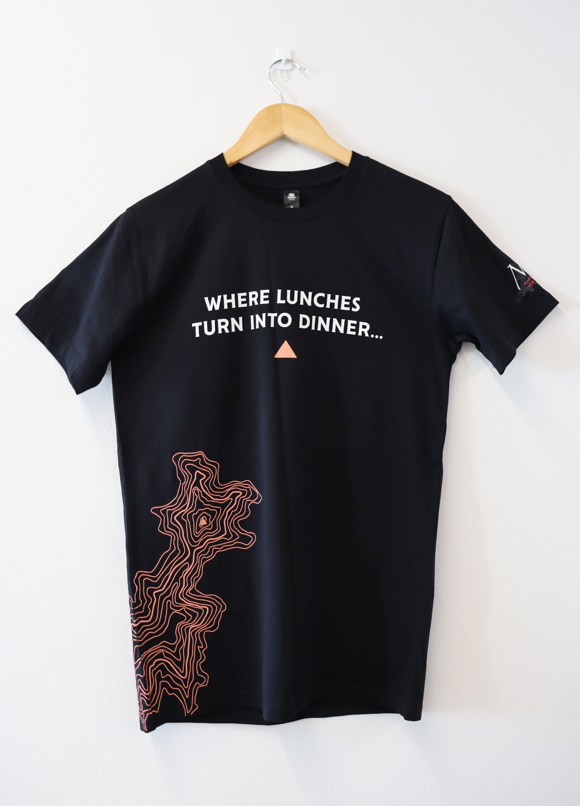 LET GO AND FEEL THE NED T-SHIRT by mariscovip, gracefully hanging on a wooden hanger against a white wall. Text reads, WHERE LUNCHES TURN INTO DINNER... with a small orange triangle. Below this, an orange contour line graphic dances elegantly on the left side of the shirt.
