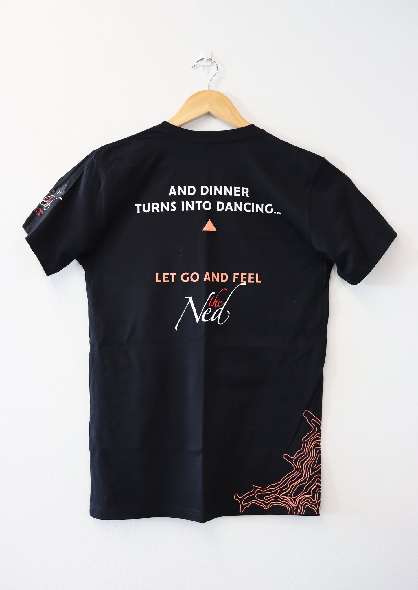 A mariscovip LET GO AND FEEL THE NED T-SHIRT in black hangs on a wooden hanger against a white wall. The back displays the phrase, "And dinner turns into dancing..." encouraging you to let go and feel the new, with decorative orange lines accentuating the bottom right.