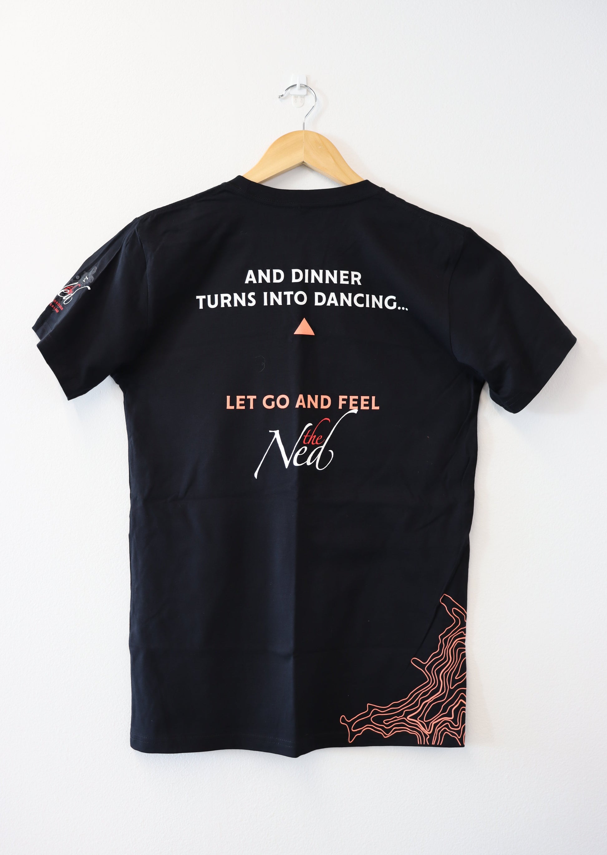A mariscovip LET GO AND FEEL THE NED T-SHIRT in black hangs on a wooden hanger against a white wall. The back displays the phrase, "And dinner turns into dancing..." encouraging you to let go and feel the new, with decorative orange lines accentuating the bottom right.