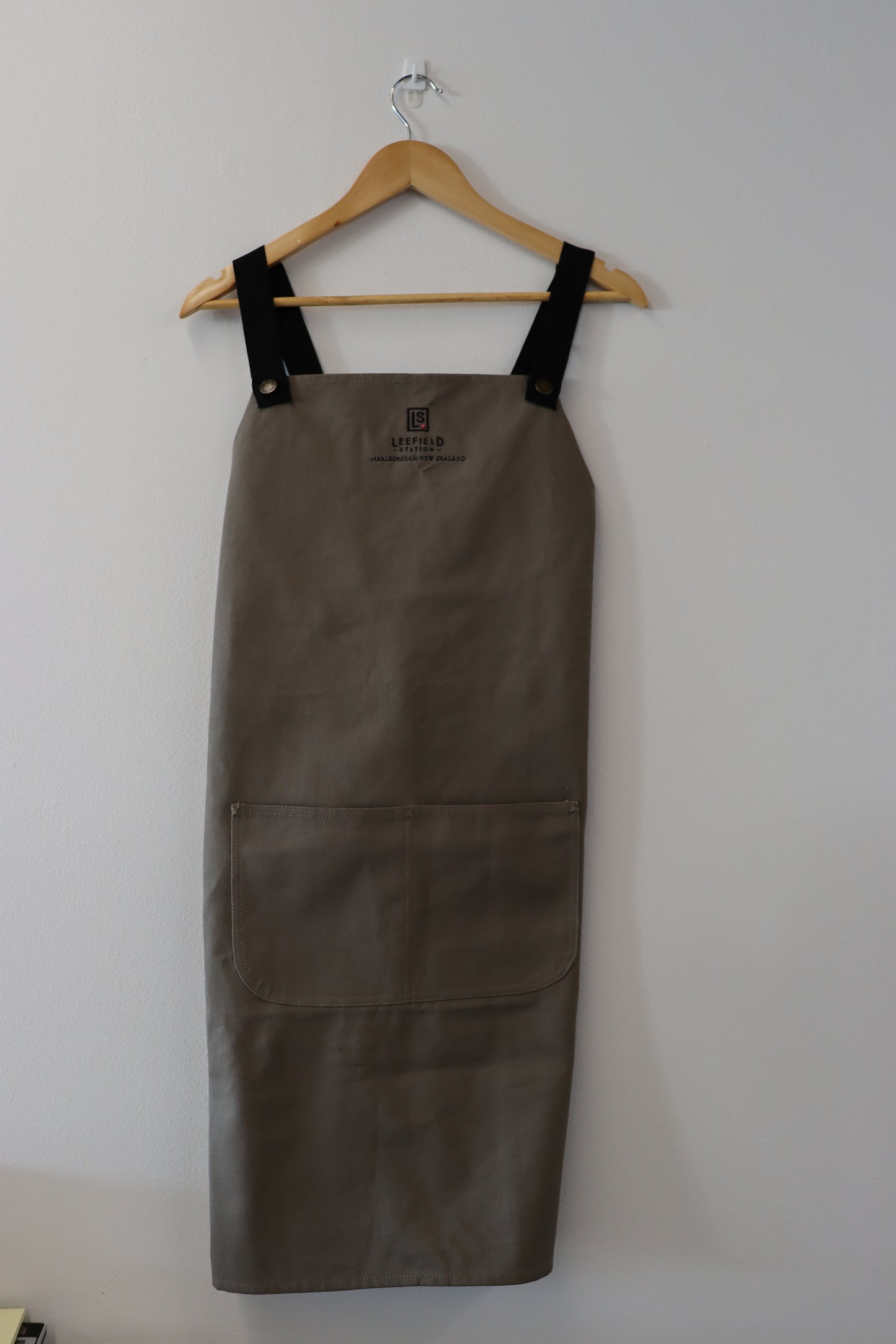 A LEEFIELD STATION APRON from Marisco Family Vineyards, featuring beige fabric with black straps, hangs on a wooden hanger against a white wall. It includes a large front pocket and displays the brand's logo with text at the top.