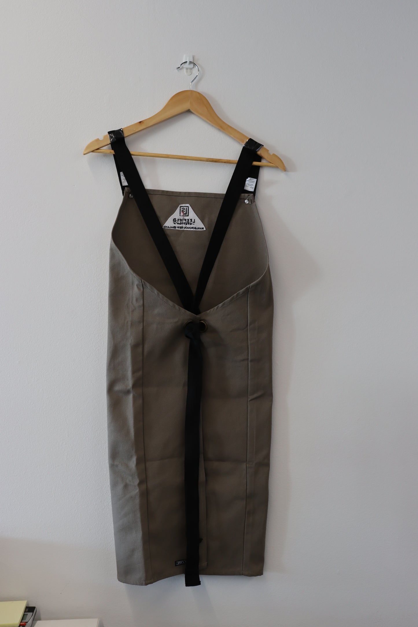 The LEEFIELD STATION APRON by Marisco Family Vineyards, featuring beige fabric with black straps, is displayed on a wooden hanger against a white wall.