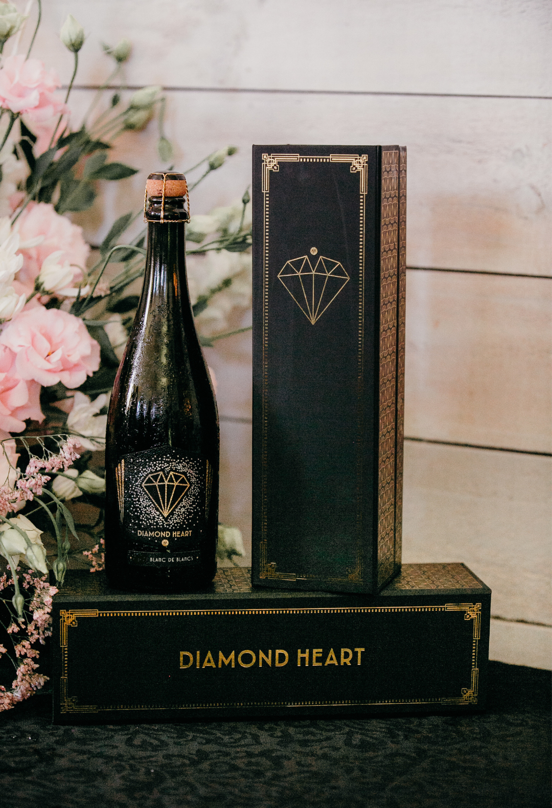 A bottle of Diamond Heart's DIAMOND HEART Wine Gift Box is elegantly displayed alongside a graceful wine gift box adorned with gold accents. The scene is beautifully enhanced with pink flowers and vibrant greenery, all set against a light wooden wall. The presentation is completed on a black lace tablecloth, exemplifying refined elegance.