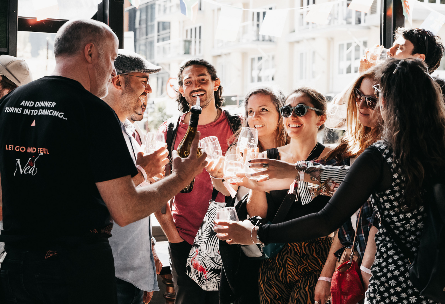 A group of people stand closely together indoors, raising their glasses for a toast. They are smiling and appear to be in a celebratory mood. Sunlight filters through the windows, creating a warm atmosphere.