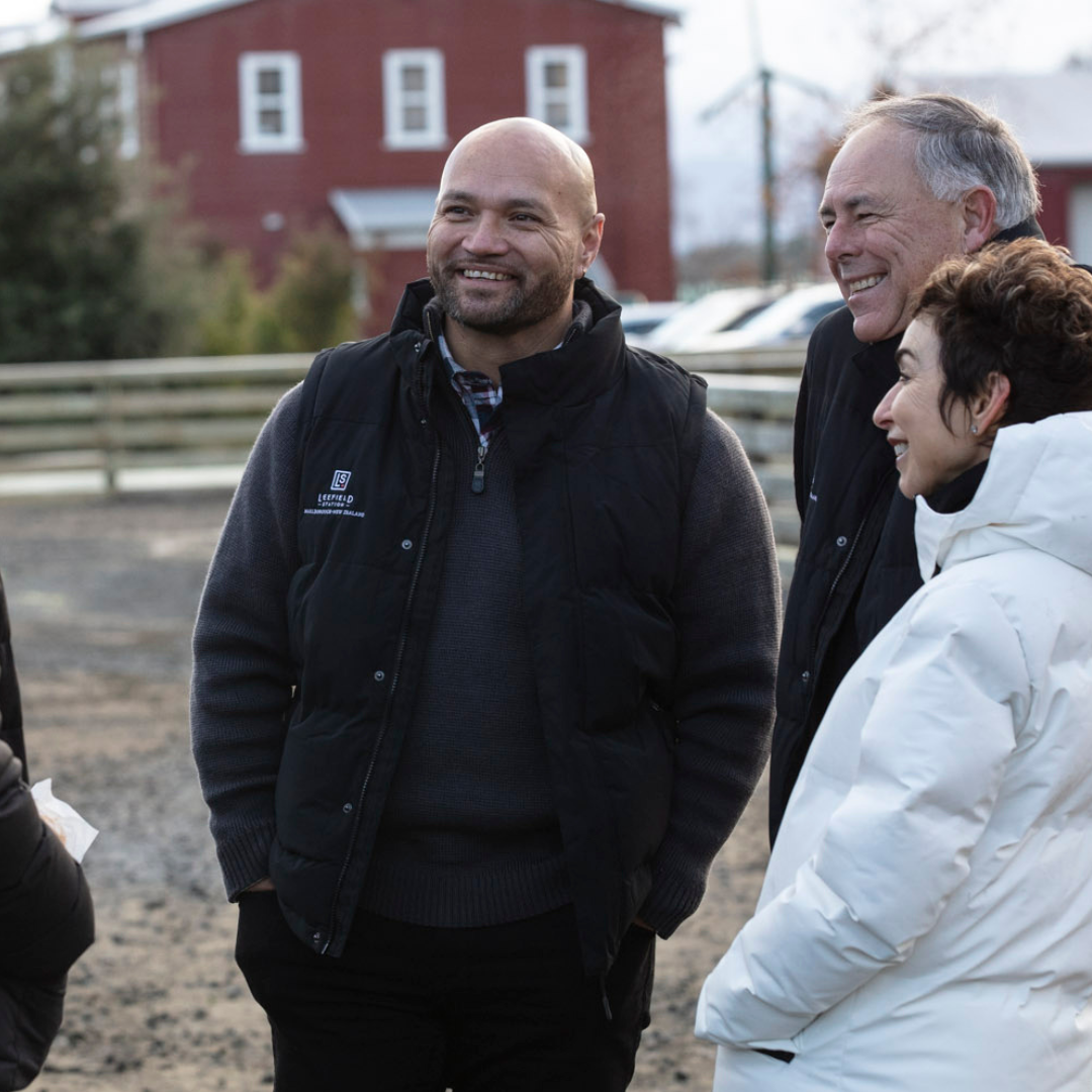 A group of four people stand outside, smiling and chatting. One person is wearing the LEEFIELD STATION PUFFER VEST from Marisco Family Vineyards. In the background, there is a red building and a wooden fence. The weather seems to be cool.