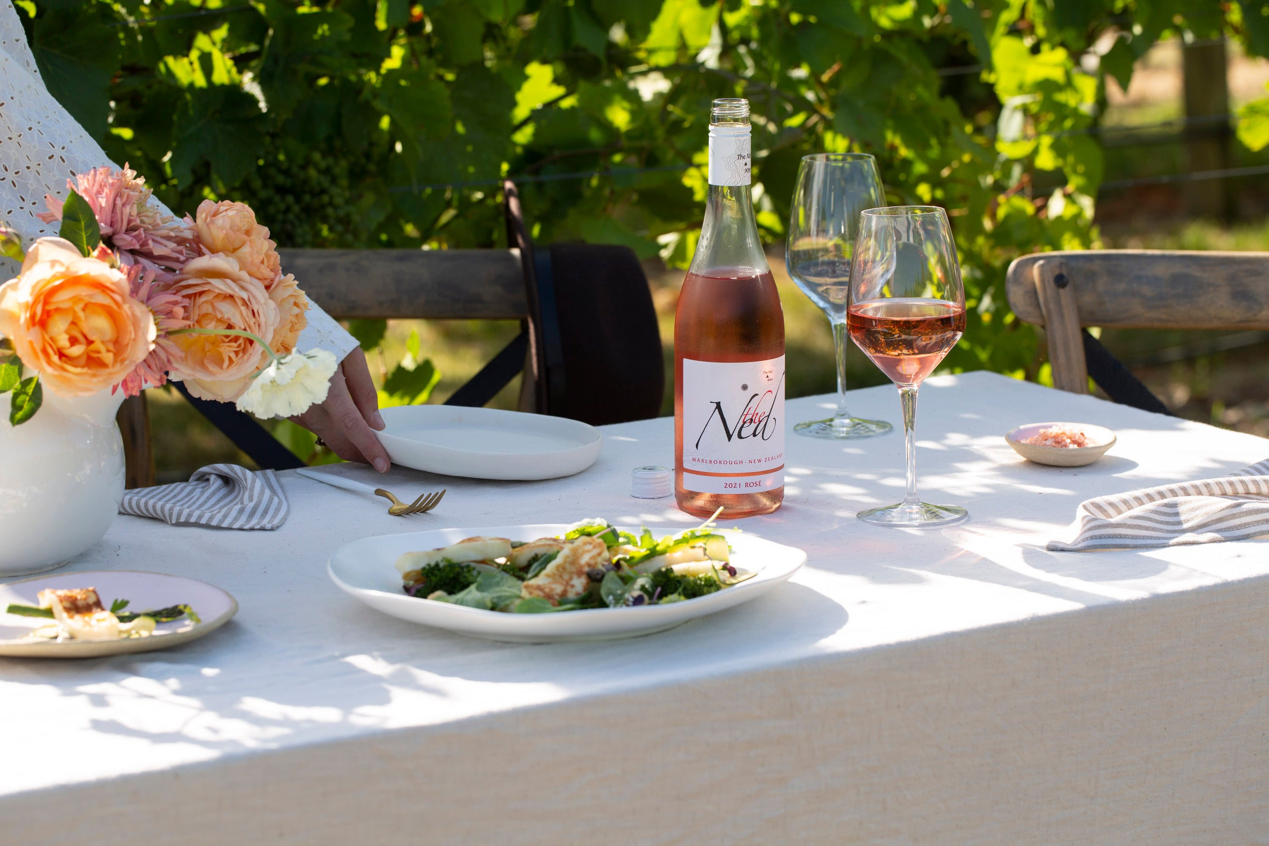 A table set outdoors with a bottle of rosé wine, two glasses filled with wine, floral centerpiece, and plates of salad. Sunlight filters through lush green grapevines in the background, creating a serene and inviting atmosphere.
