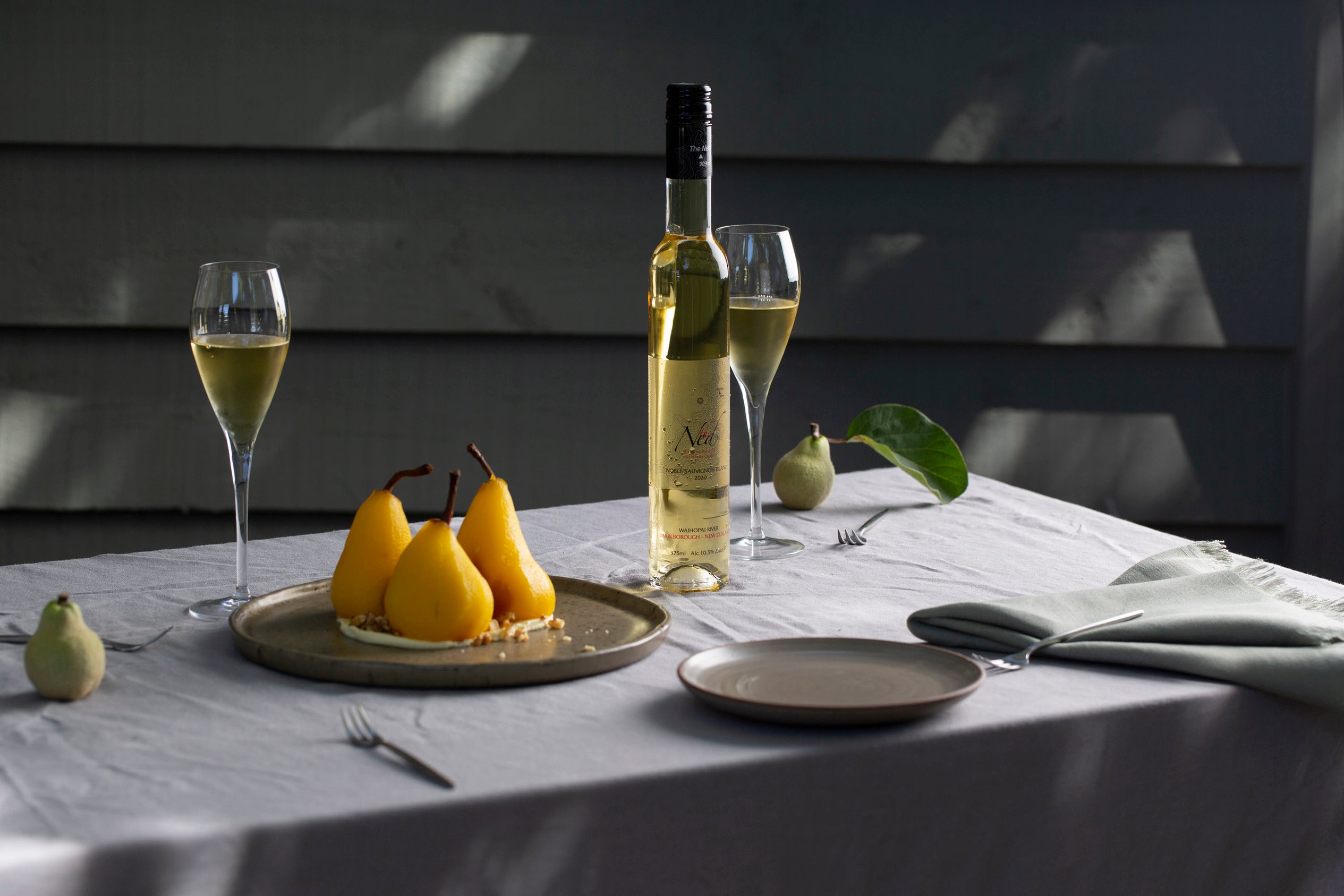 A table with a bottle of wine and two glasses filled with white wine. Three poached pears are elegantly arranged on a plate with crumble garnish. Two small, round fruits and a light gray napkin are nearby. The setting is softly lit.