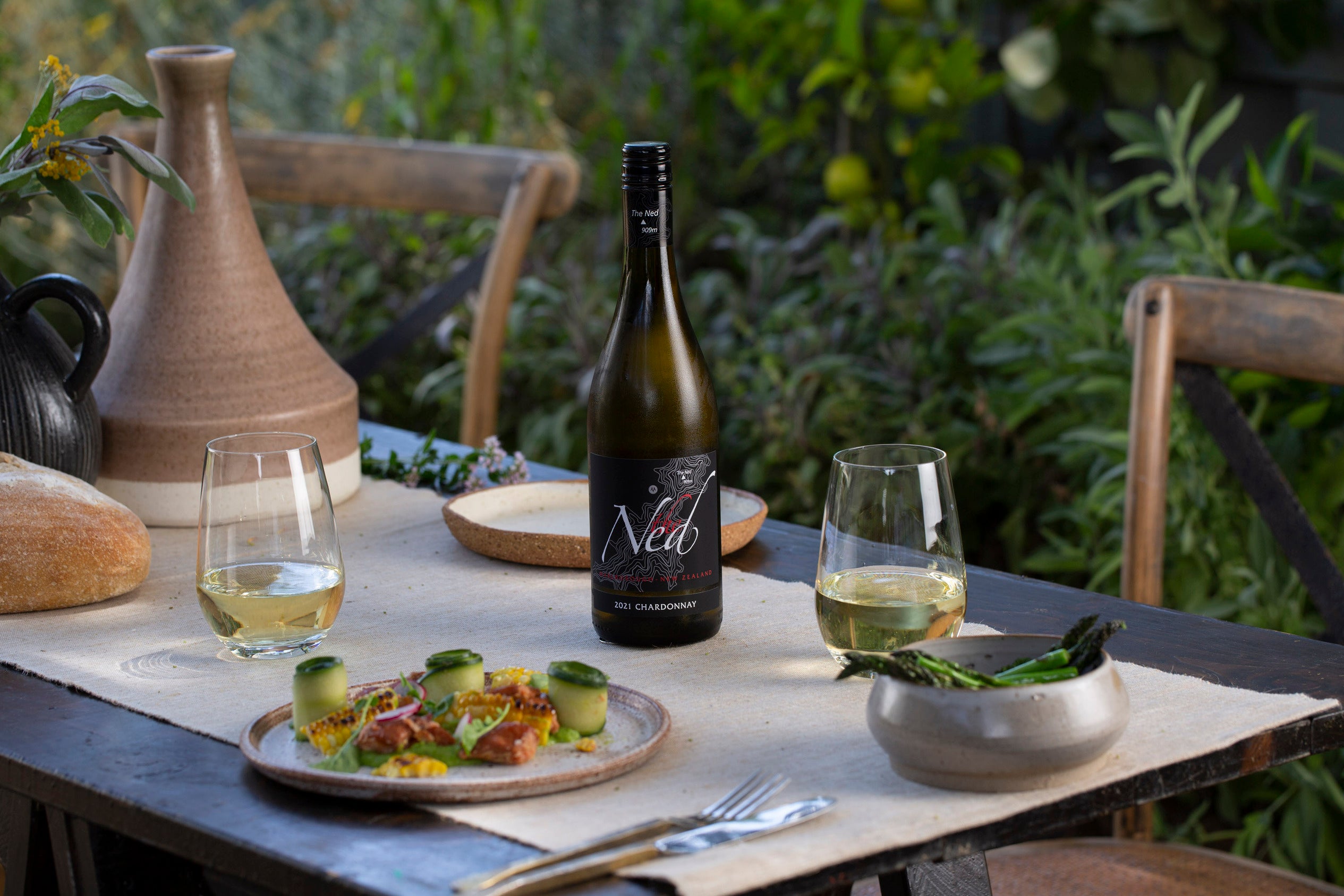 A rustic outdoor table setting features a bottle of Ned Chardonnay, two filled wine glasses, plates with appetizers, a loaf of bread, and green plants in the background, creating an inviting dining atmosphere.