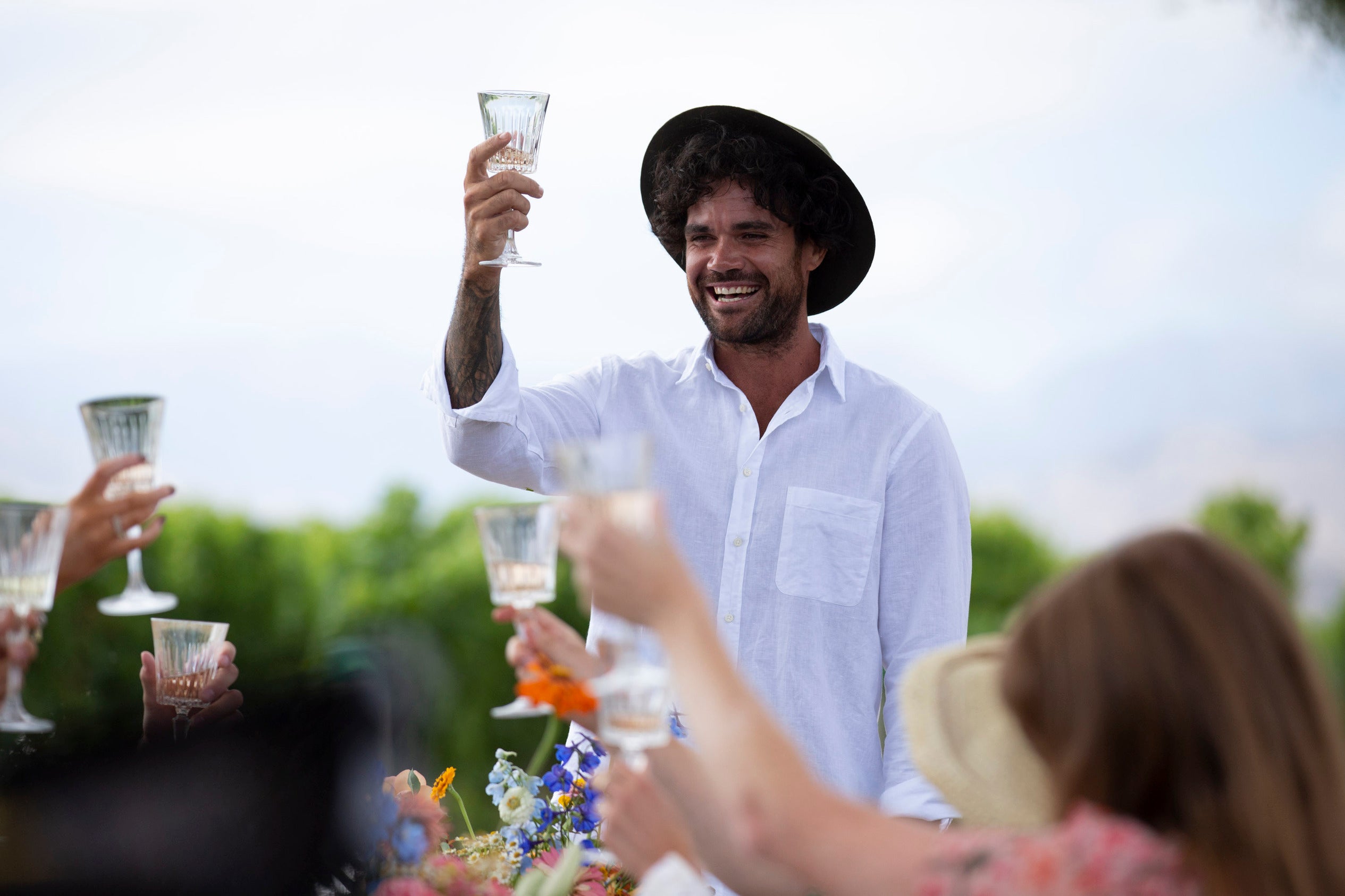 A man in a white shirt and black hat raises a glass, smiling, during an outdoor gathering. Several people around him also hold up their glasses. The background features greenery and a clear sky.