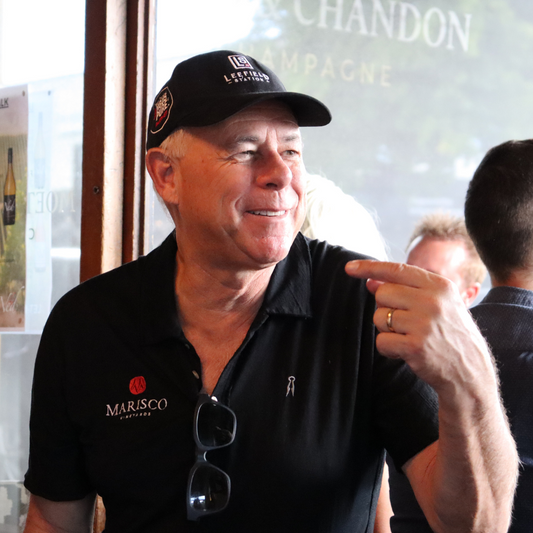 A man in a black cap labeled as the LEEFIELD STATION CAP, adorned with Marisco Family Vineyards logos on his shirt, smiles and gestures with his right hand while standing indoors. There are people in the background, and a window with partial text visible.