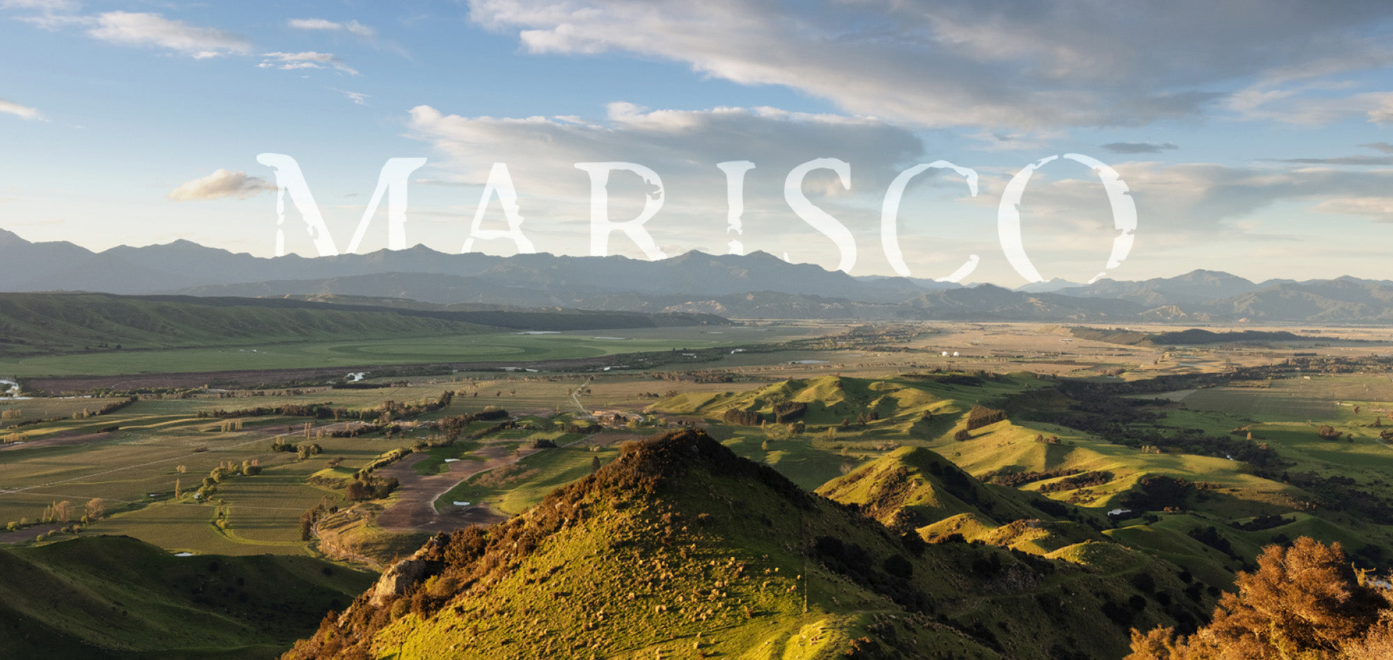 A scenic view of lush, rolling green hills under a blue sky with scattered clouds. The word MARISCO is prominently displayed in white capital letters across the image. Mountains form a distant backdrop in the landscape.