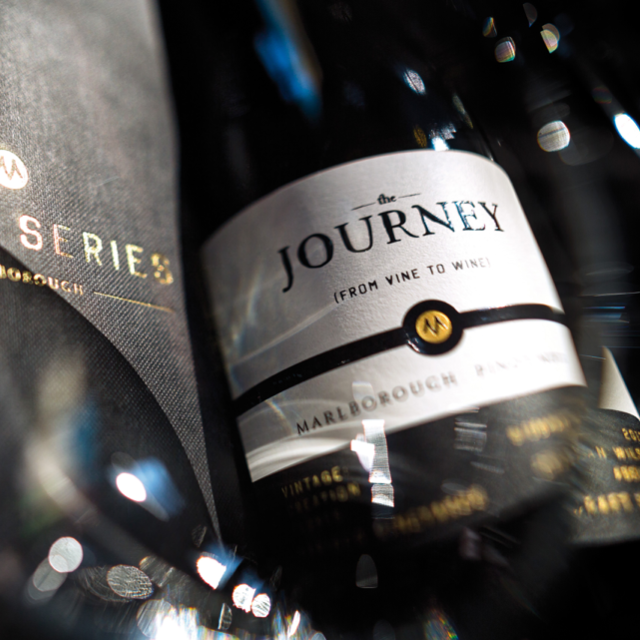 Close-up of two wine bottles. One prominently features a label reading The Journey with the tagline From Vine to Wine and mentions Marlborough Pinot Noir. The image is artistic with a soft focus and dynamic lighting.